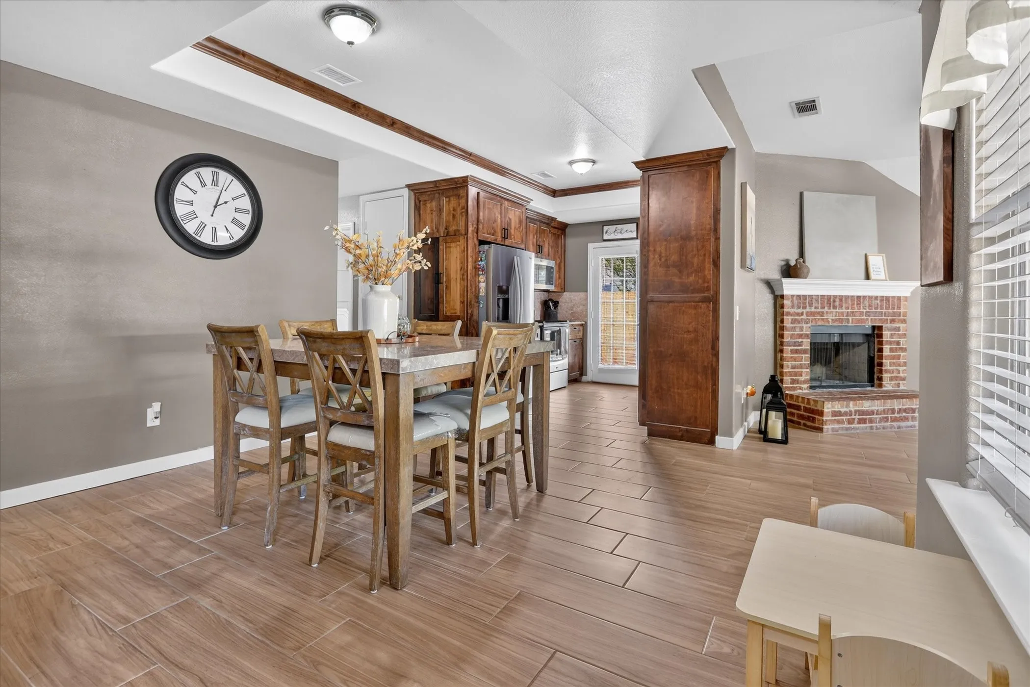 Wood-look tile flooring throughout, featuring a brick fireplace with a white mantel, and a kitchen with wood cabinetry and stainless steel appliances