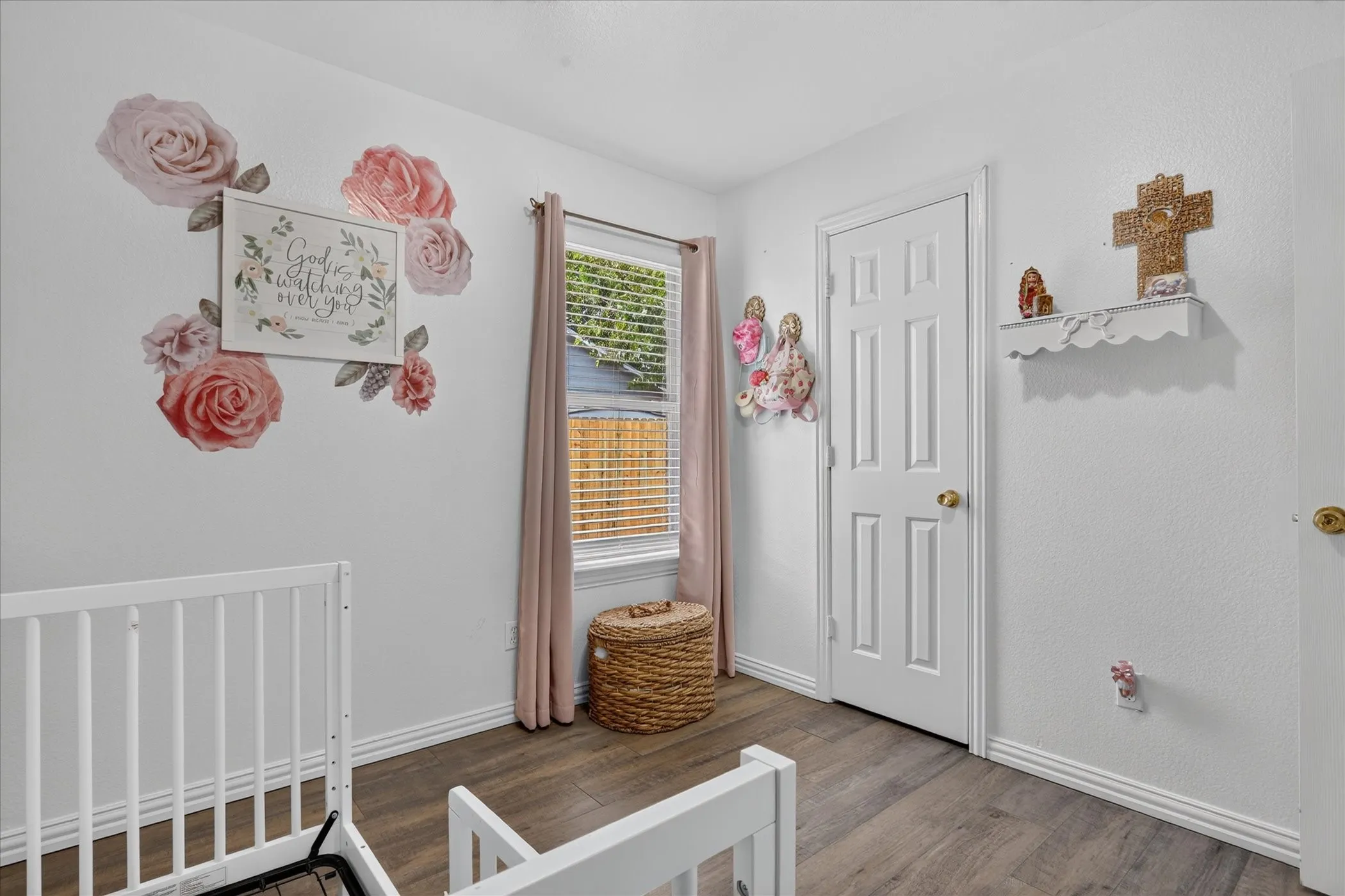 Wood-finish flooring with white baseboards, a white paneled door with a brass-finish knob, and a window with horizontal blinds