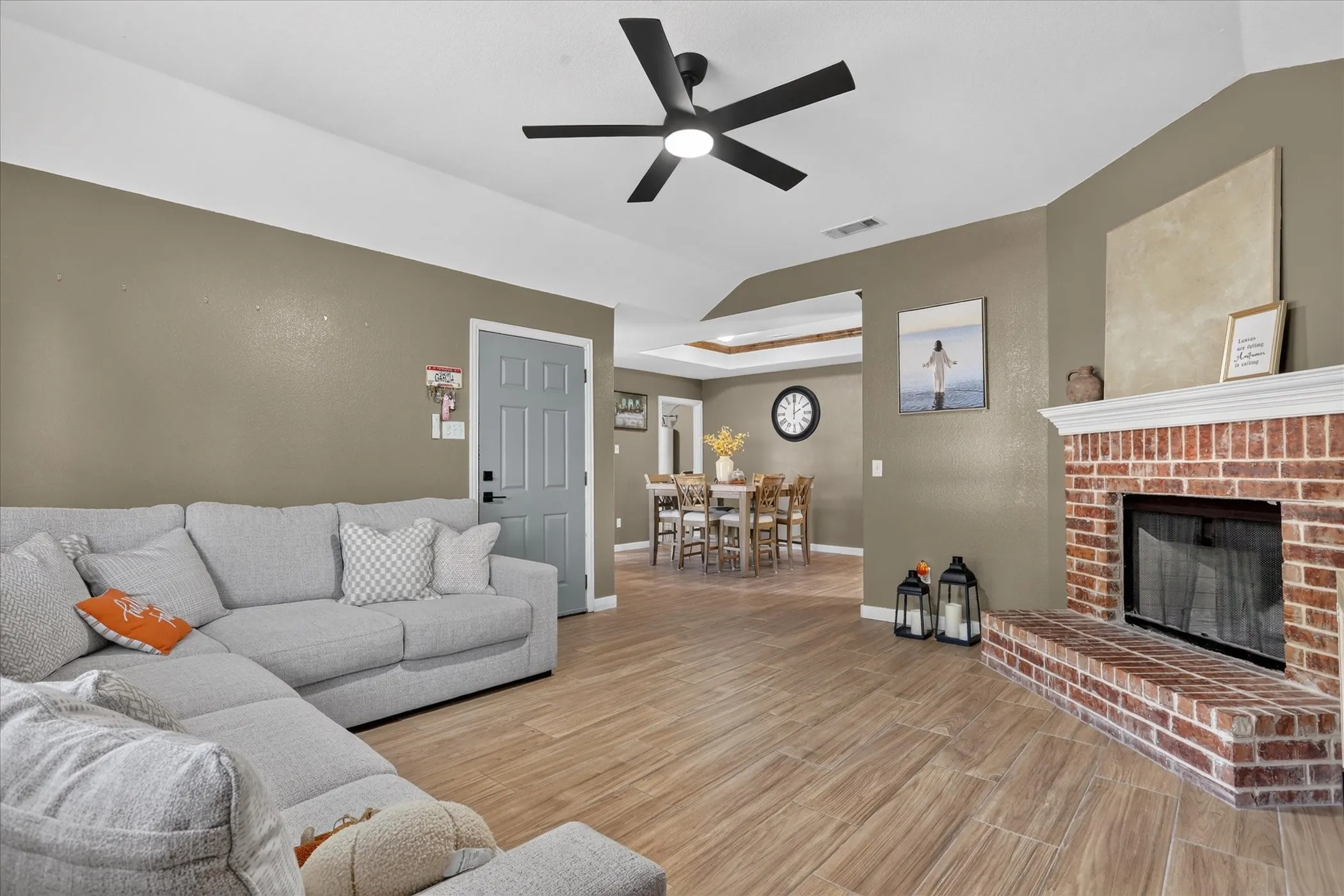 Wood-finish flooring, brick fireplace with mantel, ceiling fan with integrated lighting, and a tray ceiling detail