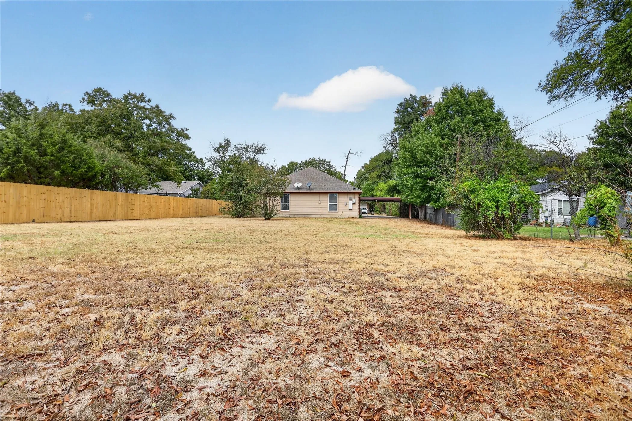 Expansive open lot with a tan brick exterior, a new wood-panel fence, a covered carport, and mature perimeter trees