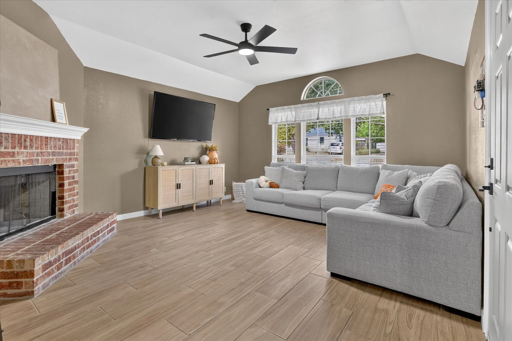 Vaulted ceiling with a black ceiling fan, brick fireplace with a white mantel, wood-finish tile flooring, and a large window with an arched transom
