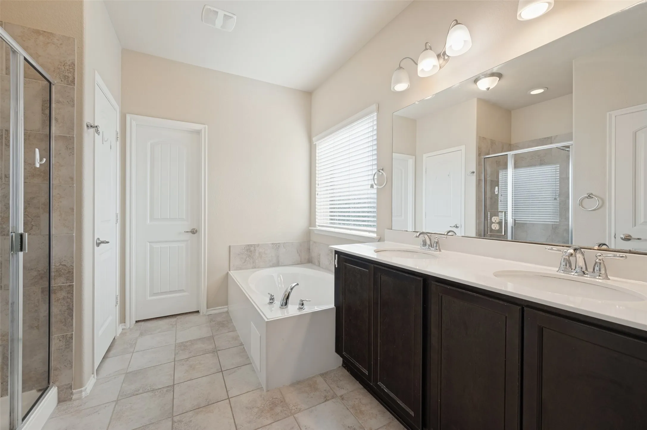 Dual vanity with dark wood-finish cabinetry and white countertops, a built-in soaking tub, a separate glass-enclosed shower, and neutral tile flooring