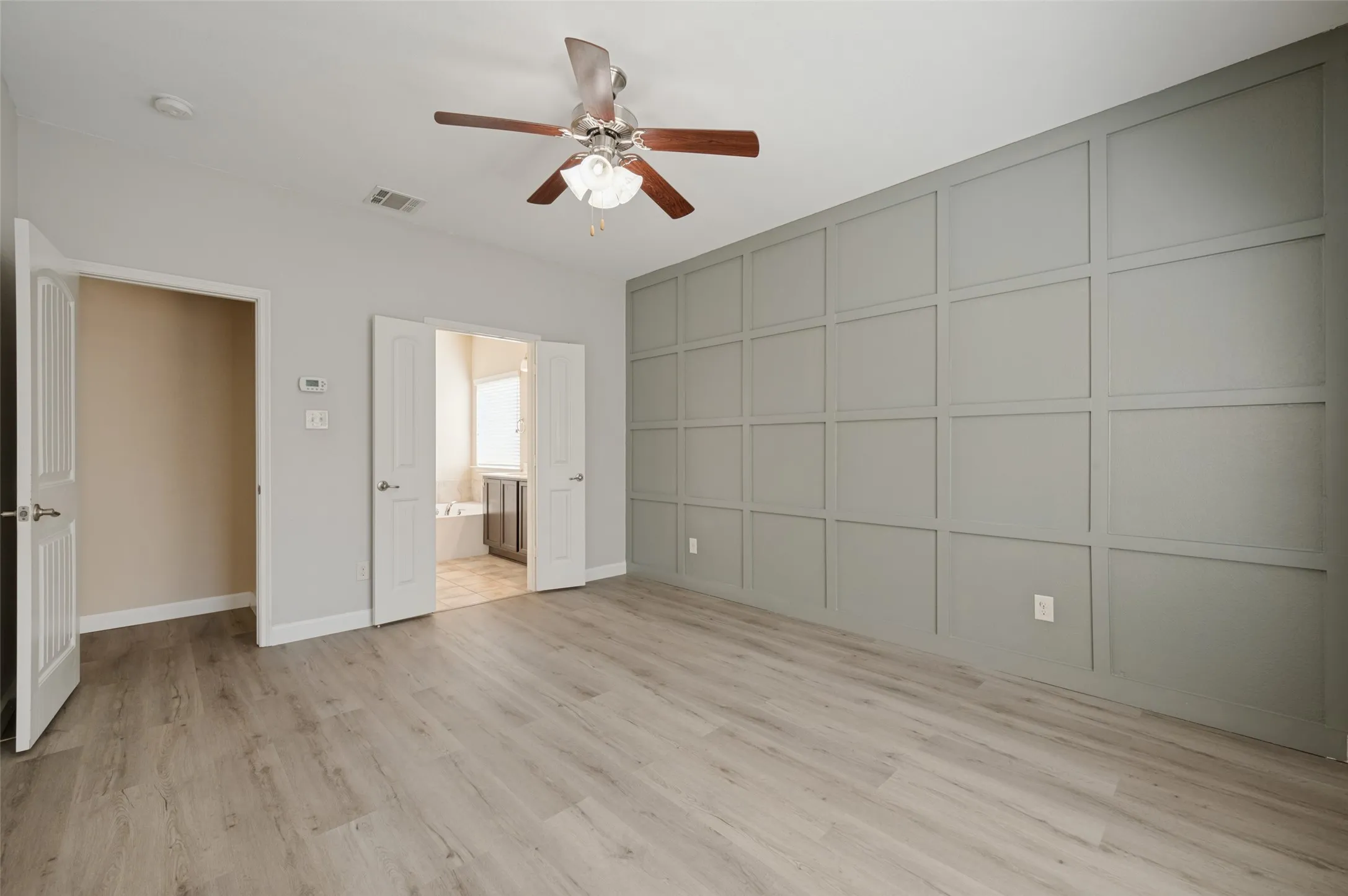 Wood-finish flooring, accent wall with panel detailing, ceiling fan with light fixture, double doors, and a built-in bathtub