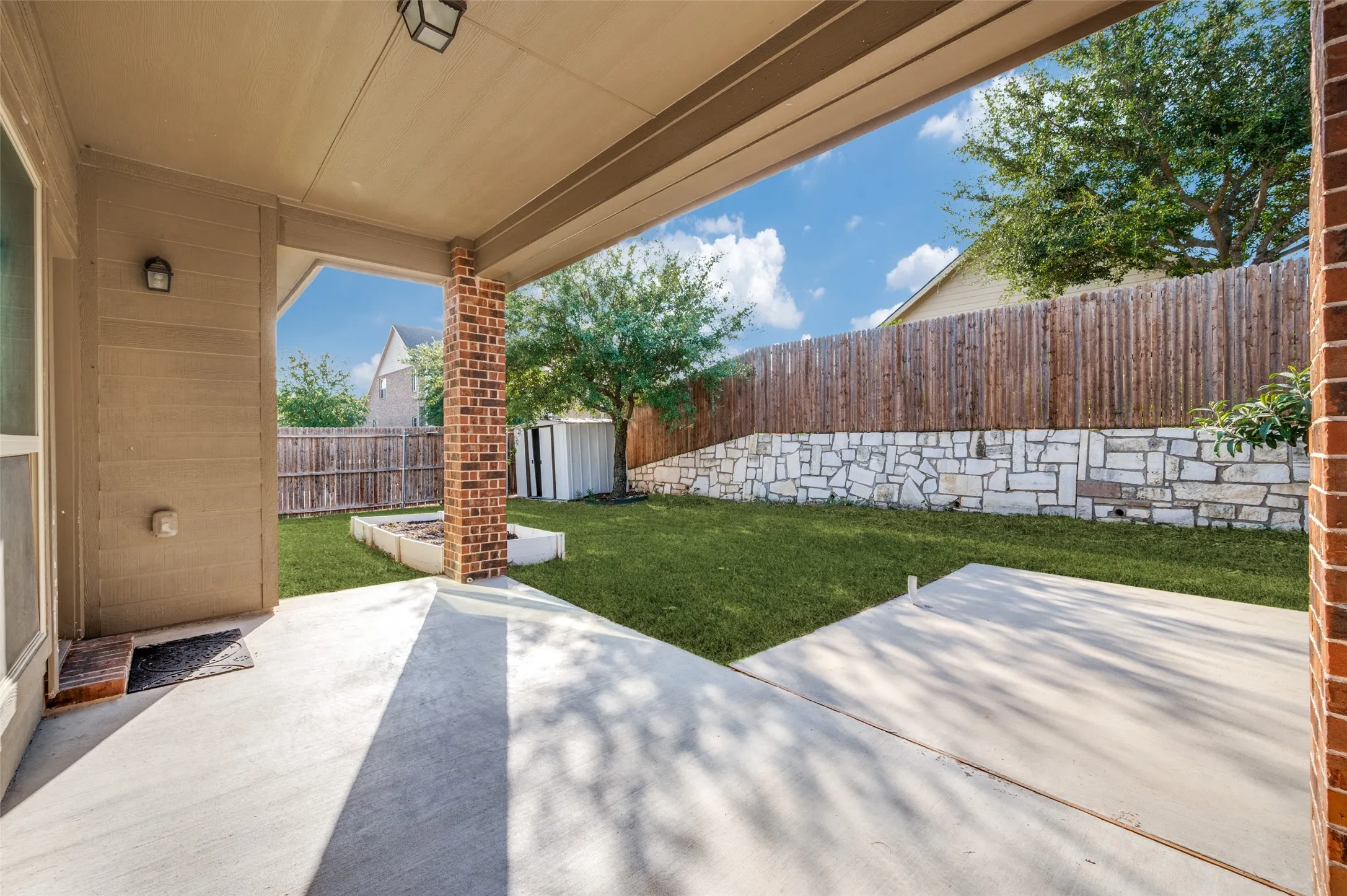 Covered patio with a concrete surface, brick columns, and a landscaped backyard featuring a stone retaining wall and a wooden privacy fence