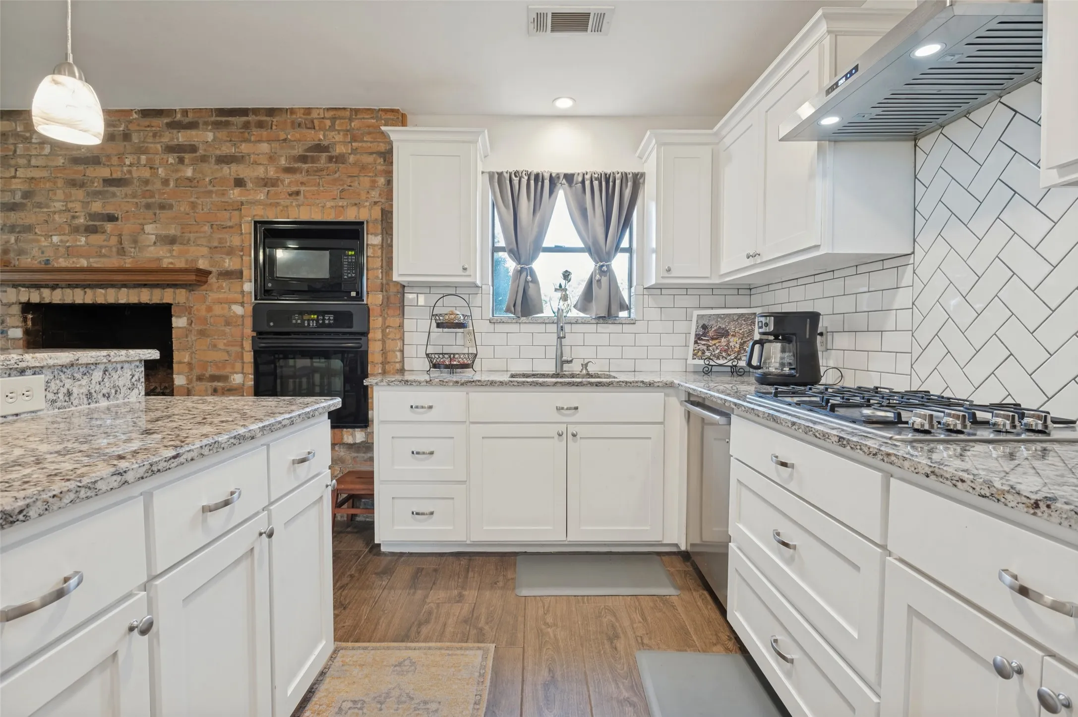 Kitchen featuring white cabinetry, light-toned countertops, a herringbone tile backsplash, wood-finish flooring, and a brick accent wall with a built-in oven and microwave