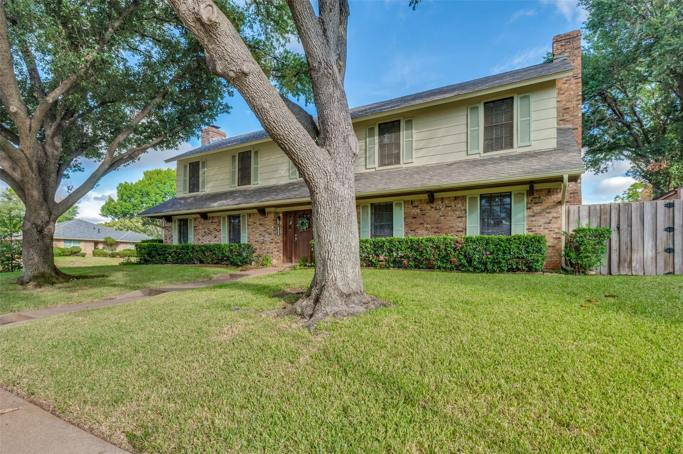 Traditional two-story exterior with brick and siding facade, featuring a prominent chimney, established trees, and a privacy fence