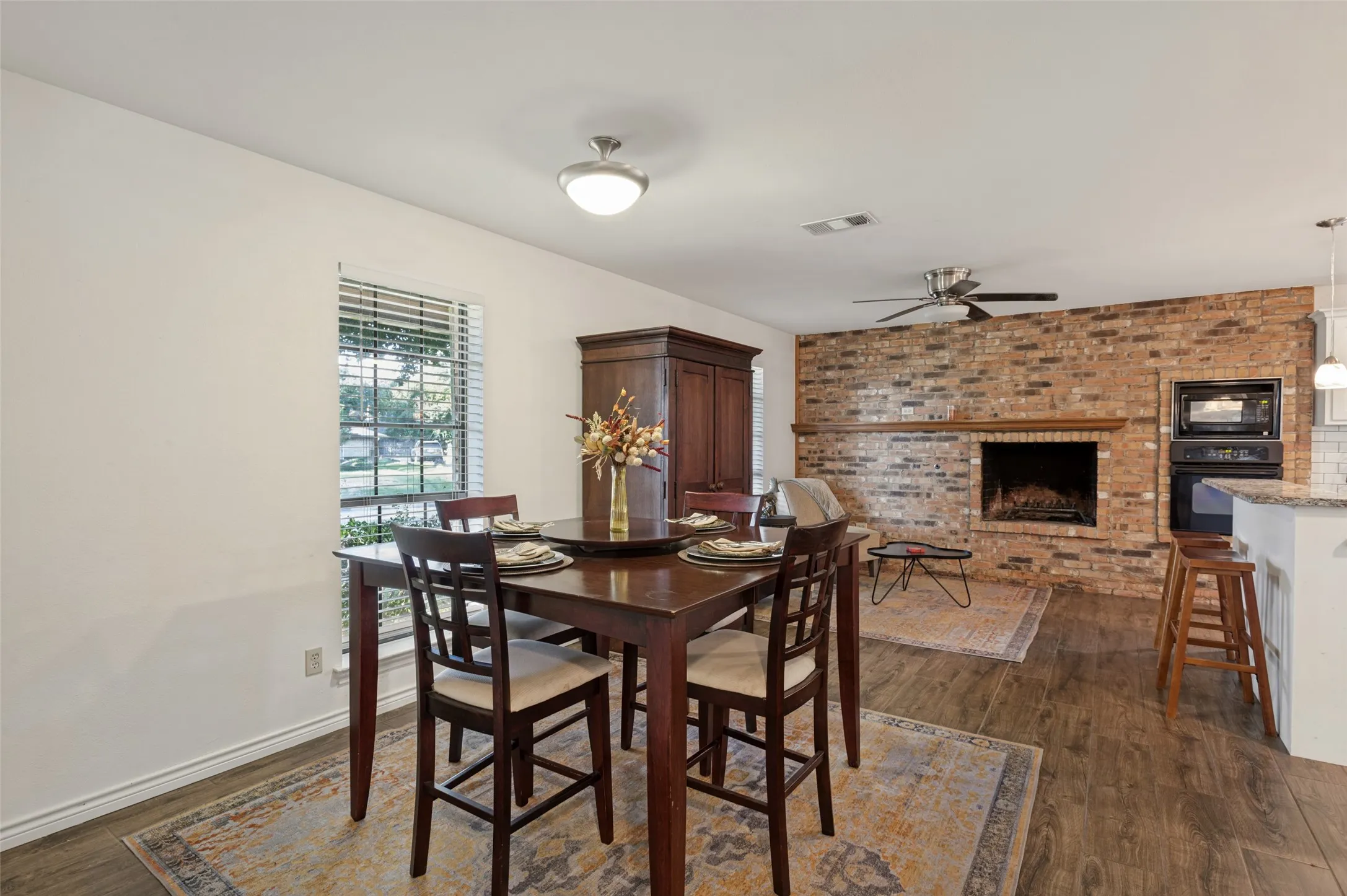 Wood-finish flooring, brick fireplace with mantel, white walls, built-in oven and microwave, and ceiling fan