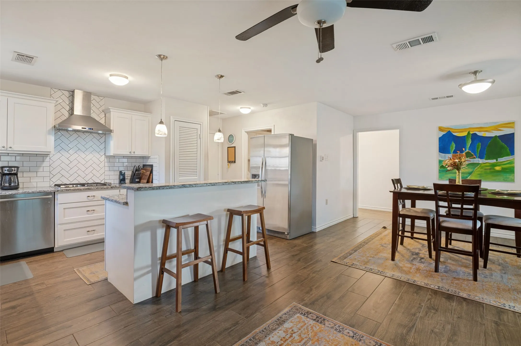 Kitchen and dining area featuring wood-finish flooring, white cabinetry, stainless steel appliances, a white subway tile backsplash, and a breakfast bar with pendant lighting