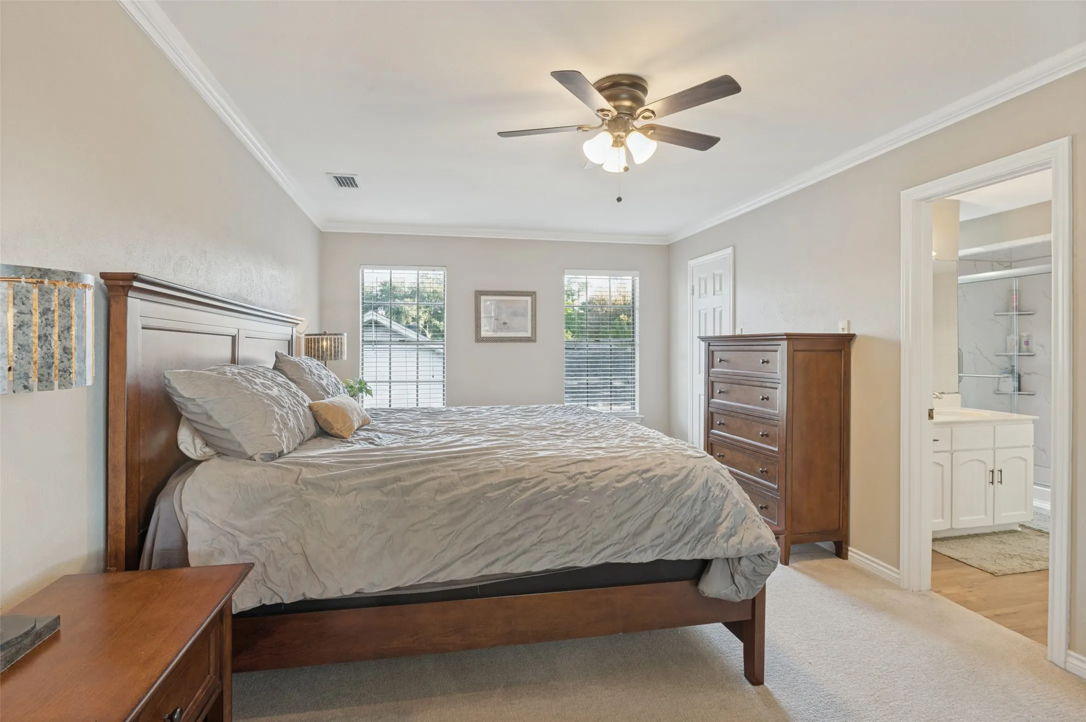 Neutral wall finishes, crown molding, ceiling fan with integrated lighting, natural light from two windows, and an en-suite bathroom with a white vanity and shower