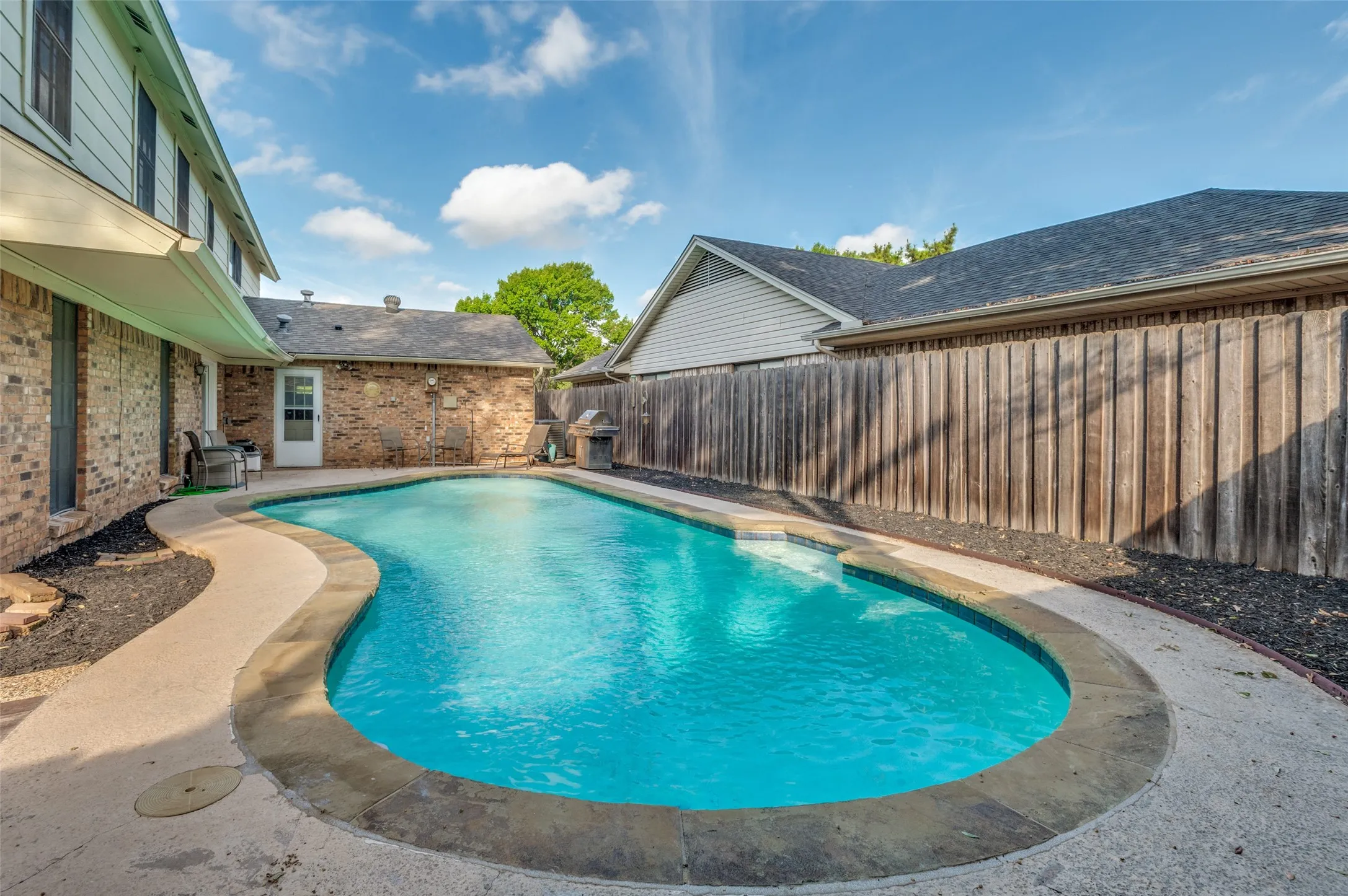 Curved swimming pool with concrete decking, brick exterior, and wood fencing