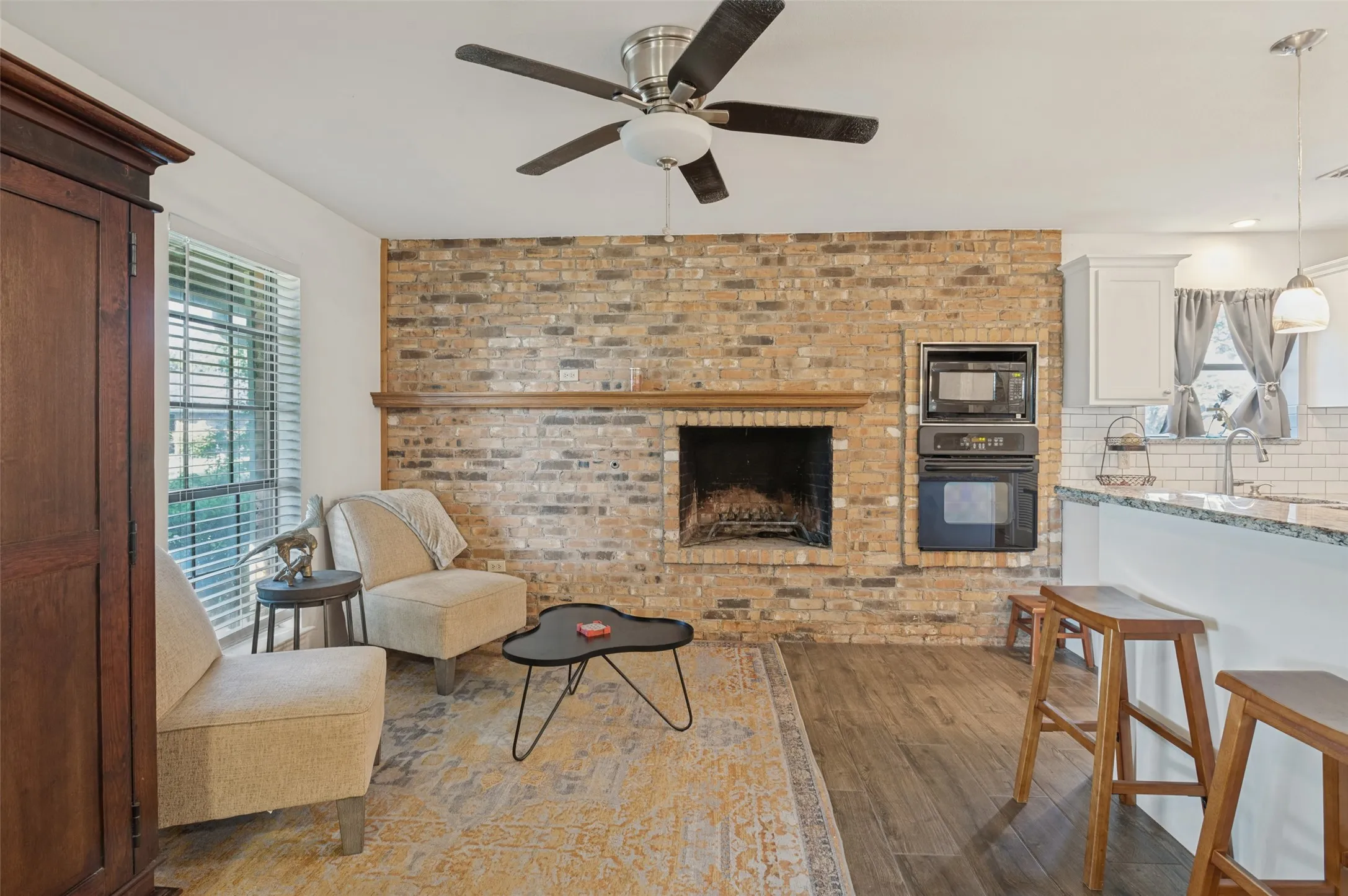 Brick-surround fireplace with a wood mantle, built-in wall oven and microwave, wood-finish flooring, and a ceiling fan