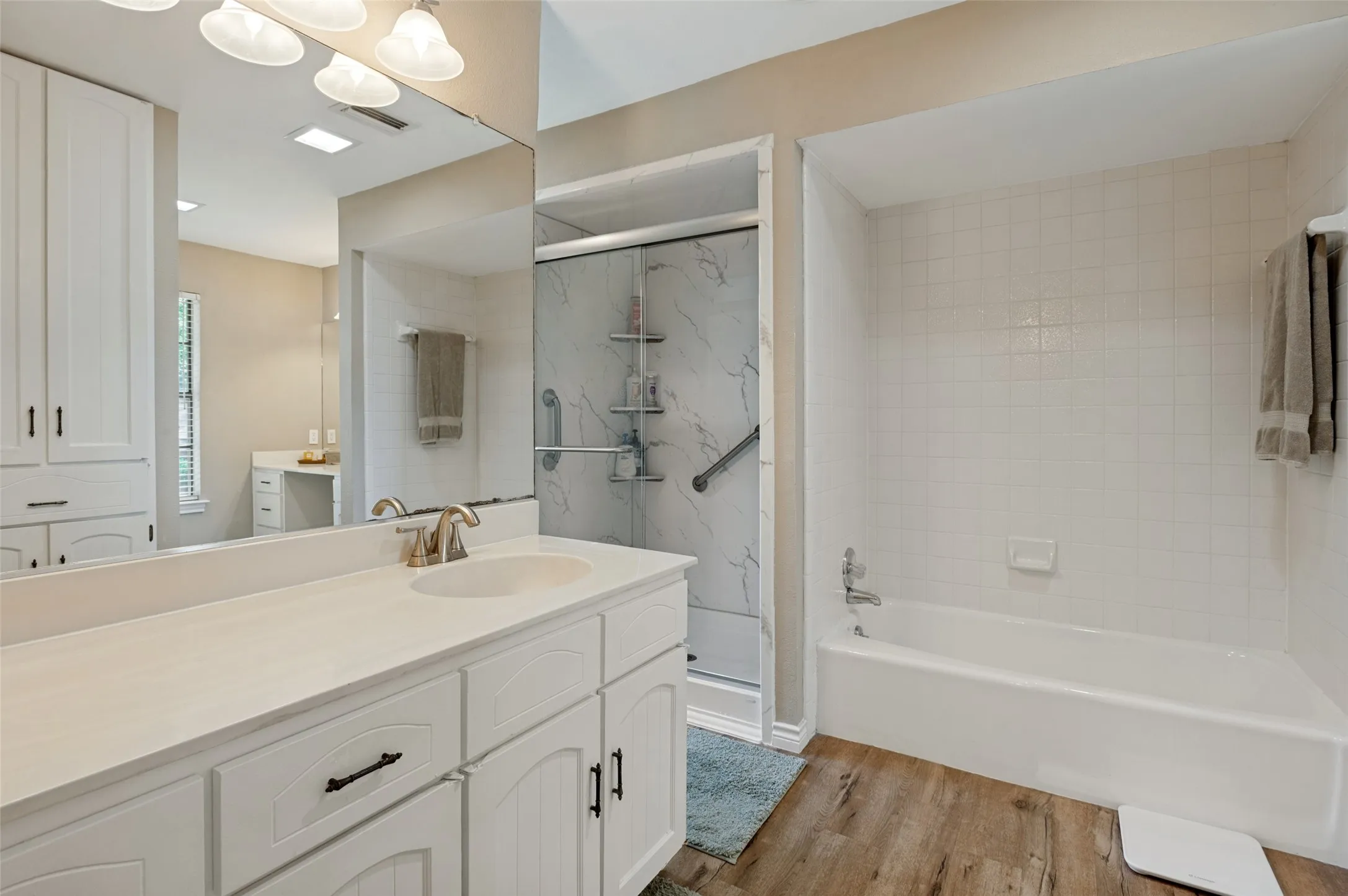 White vanity with an integrated sink and brushed nickel faucet, large wall mirror, wood-finish flooring, and a shower-tub combination with white tile surround