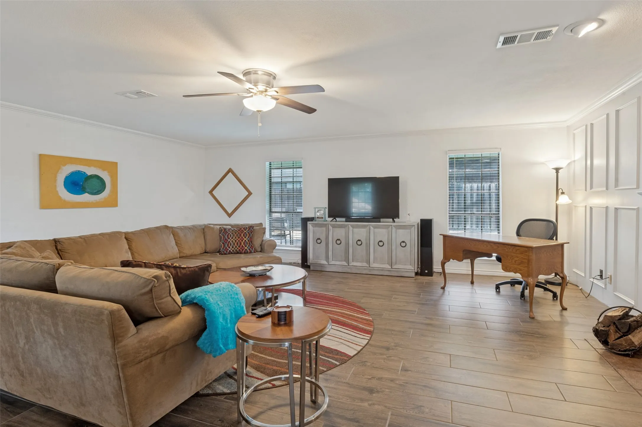 Wood-finish flooring throughout, ceiling fan with integrated lighting, multiple windows with blinds, built-in panel accent wall, and crown molding