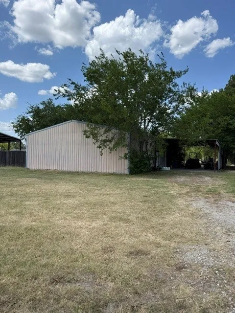 Metal utility building with an attached covered structure, expansive grassy area, and mature trees