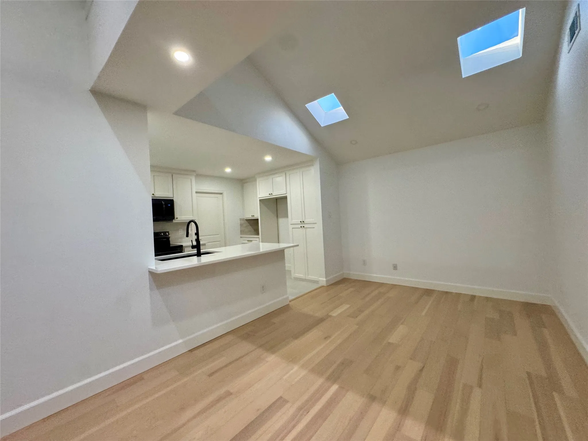 Vaulted ceiling with multiple skylights, recessed lighting, wood-finish flooring, and a kitchen featuring white cabinetry, a black faucet, and built-in appliances