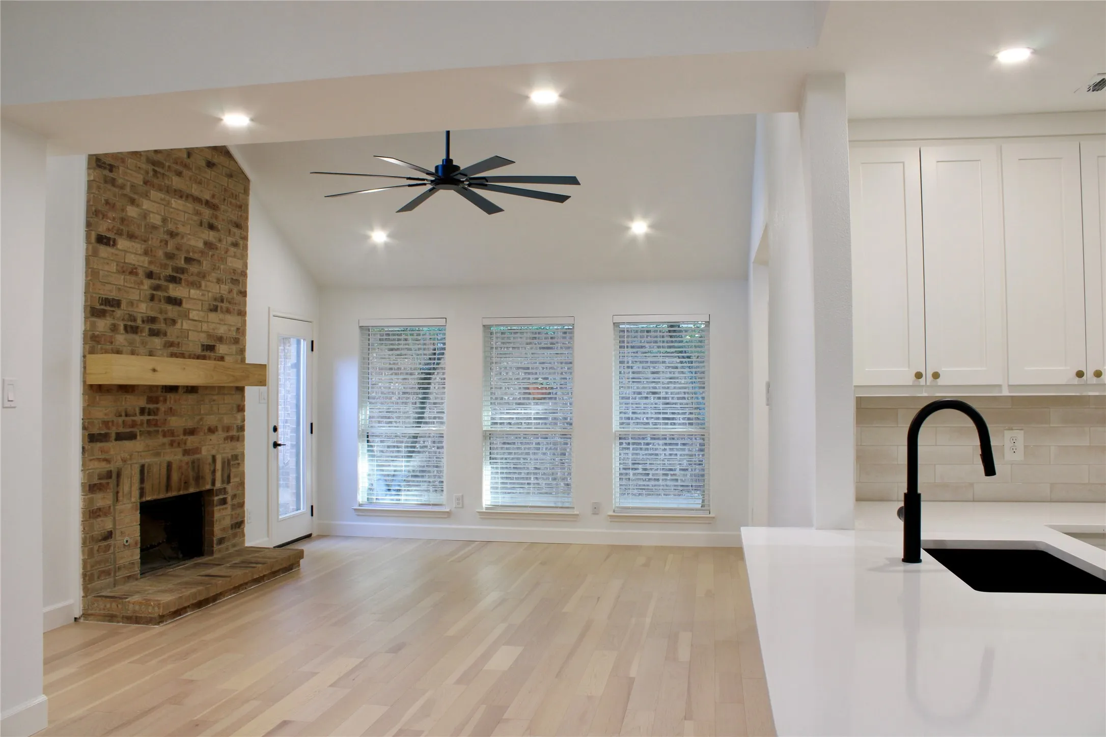 Vaulted ceiling with a ceiling fan, brick fireplace with a wood mantle, light wood-finish flooring, three windows, and white kitchen cabinetry with a matte black faucet