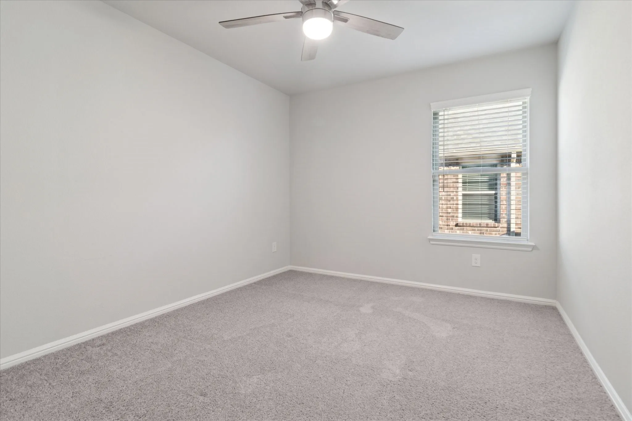 Light gray carpet flooring, white painted walls, single window with blinds, and a ceiling fan with integrated lighting