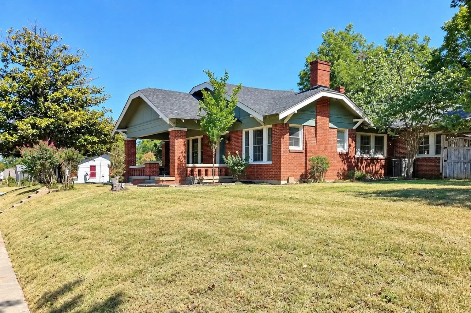 Wide-angle exterior view showing the home’s red-brick construction, multiple rooflines, chimney, porch, large front lawn, and mature trees.