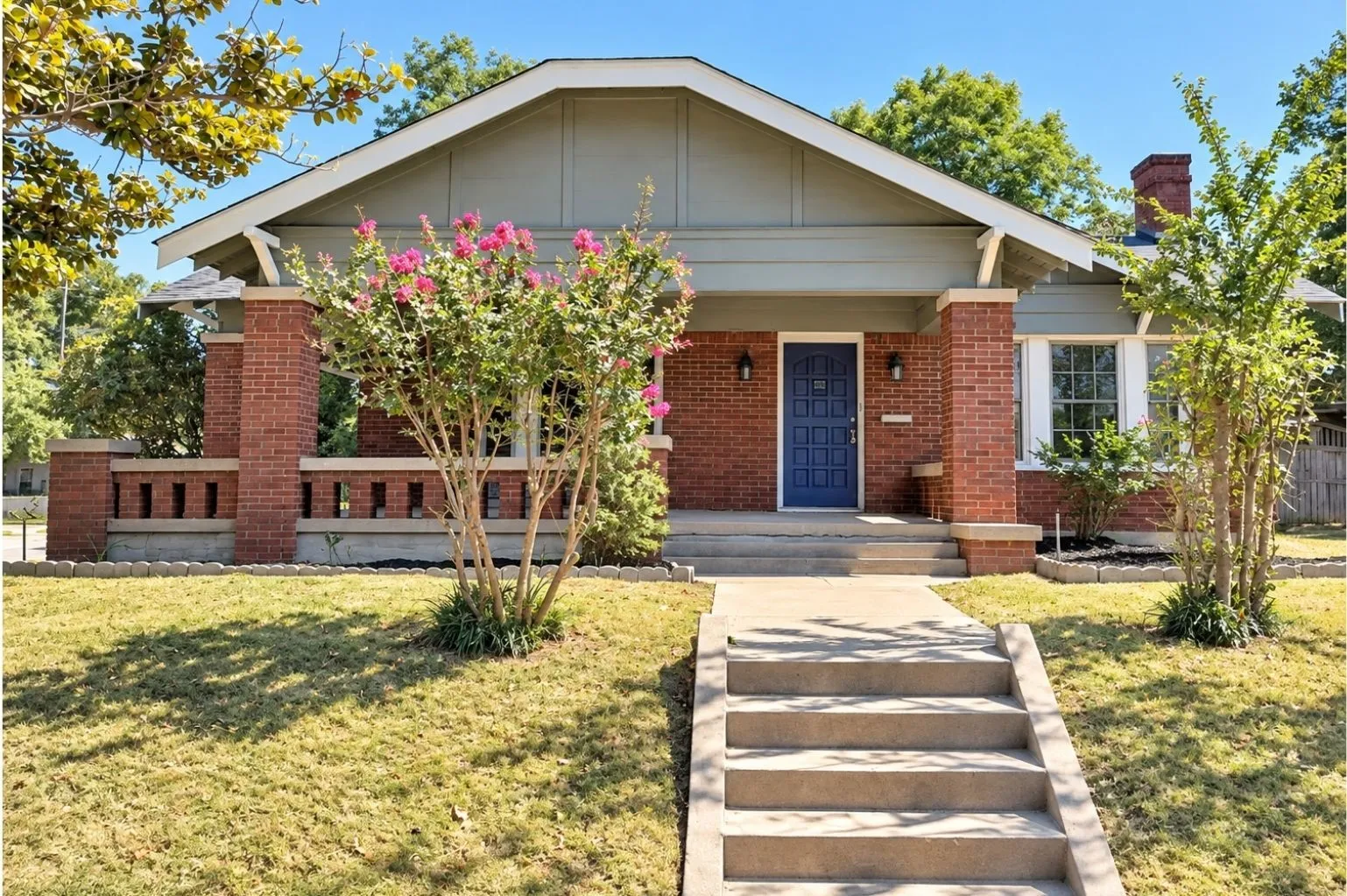 Straight-on exterior view highlighting the home’s covered porch, blue front door, brick columns, gabled roofline, and front landscaping.