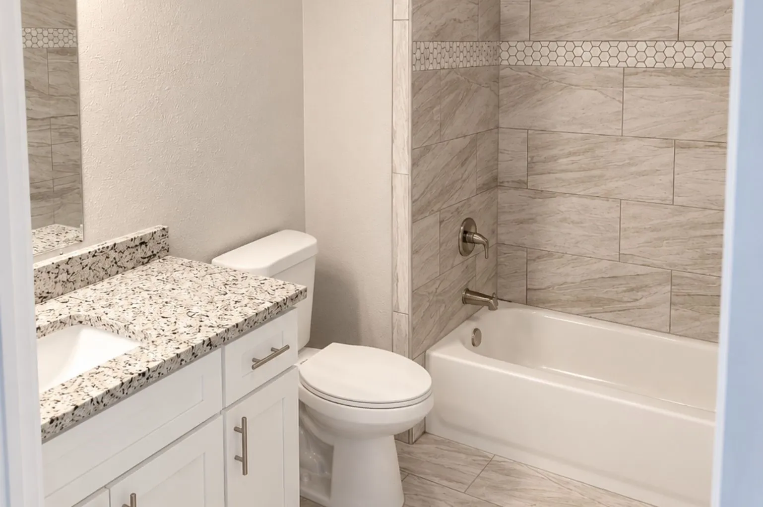 Updated bathroom featuring a white vanity, granite countertop, toilet, tub-shower combination, marble-look tile surround, and mosaic accent strip.
