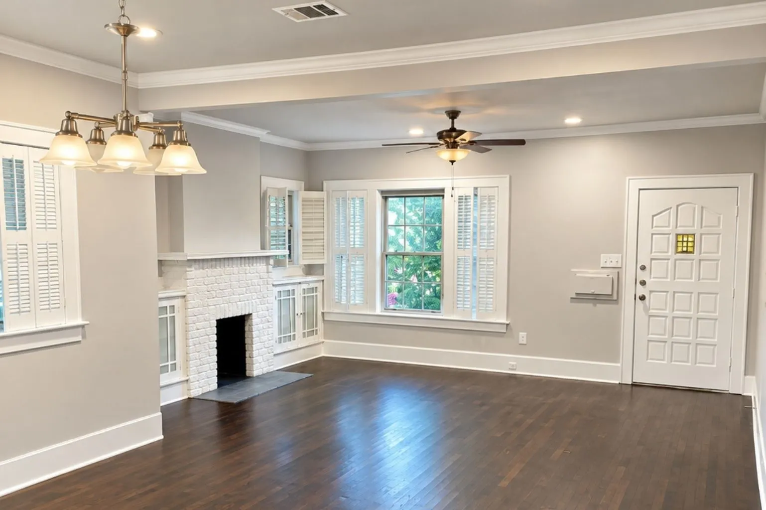Bright, spacious living area featuring dark hardwood floors, white brick fireplace, built-in cabinetry, plantation shutters, ceiling fan, and front entry.