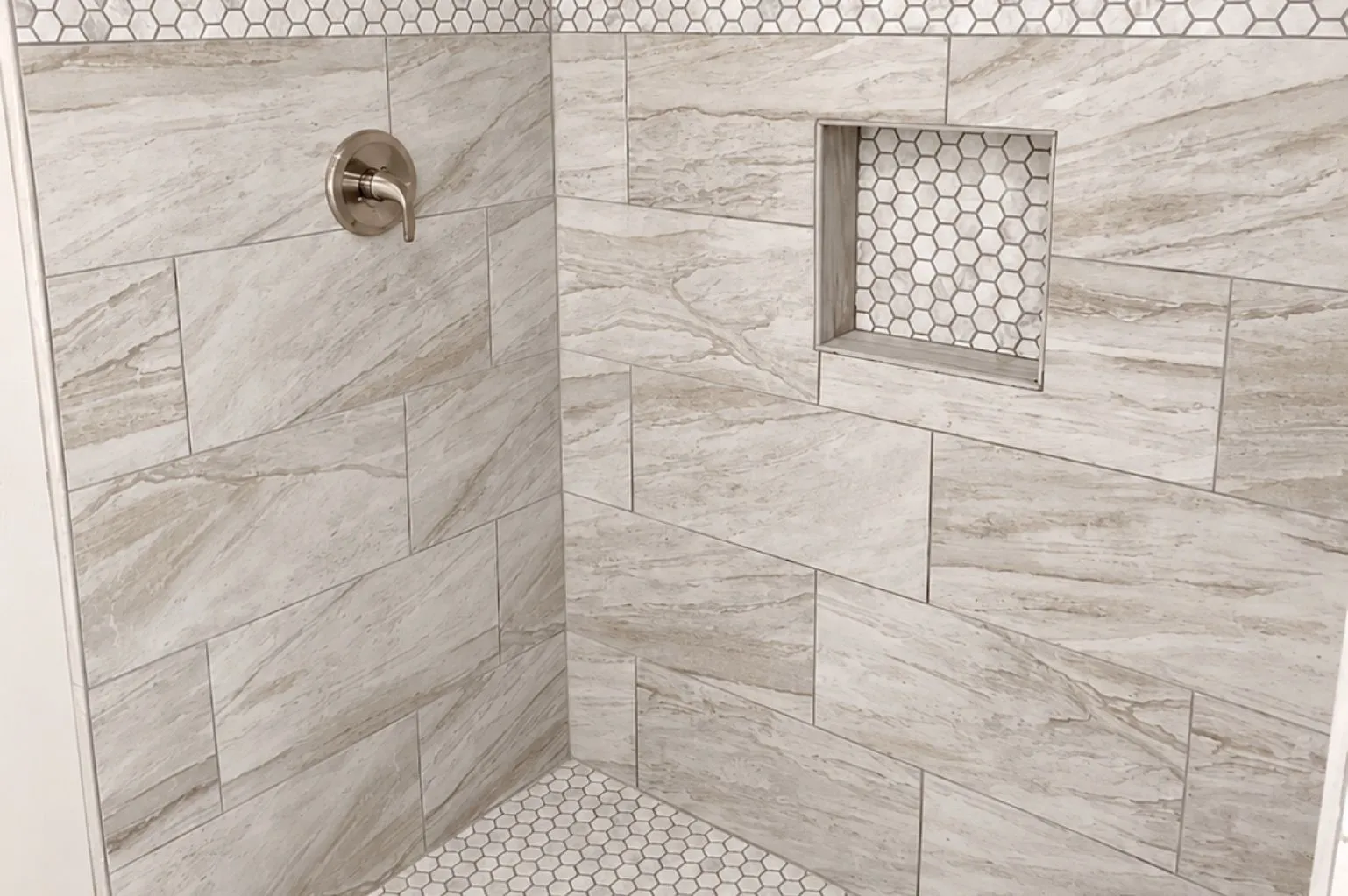 Close-up view of the tiled walk-in shower featuring marble-look wall tile, hexagon mosaic accents, recessed niche, and brushed-nickel fixtures.