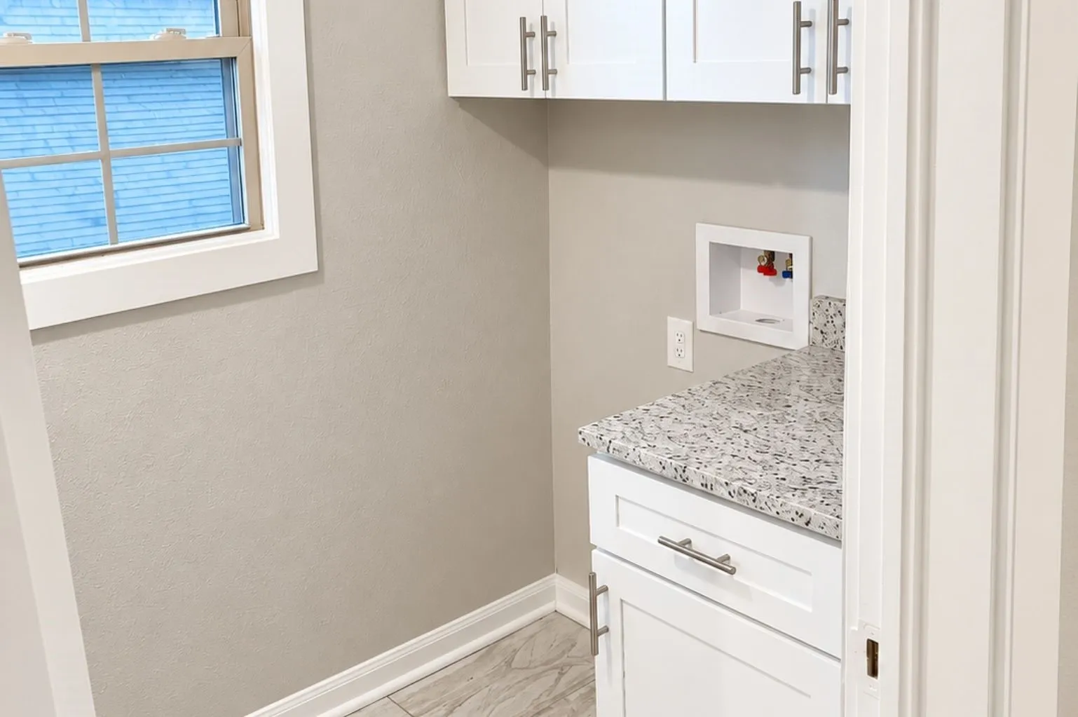 Compact laundry and utility room featuring upper and lower white cabinetry, granite counter, washer hookups, window, and light tile flooring.