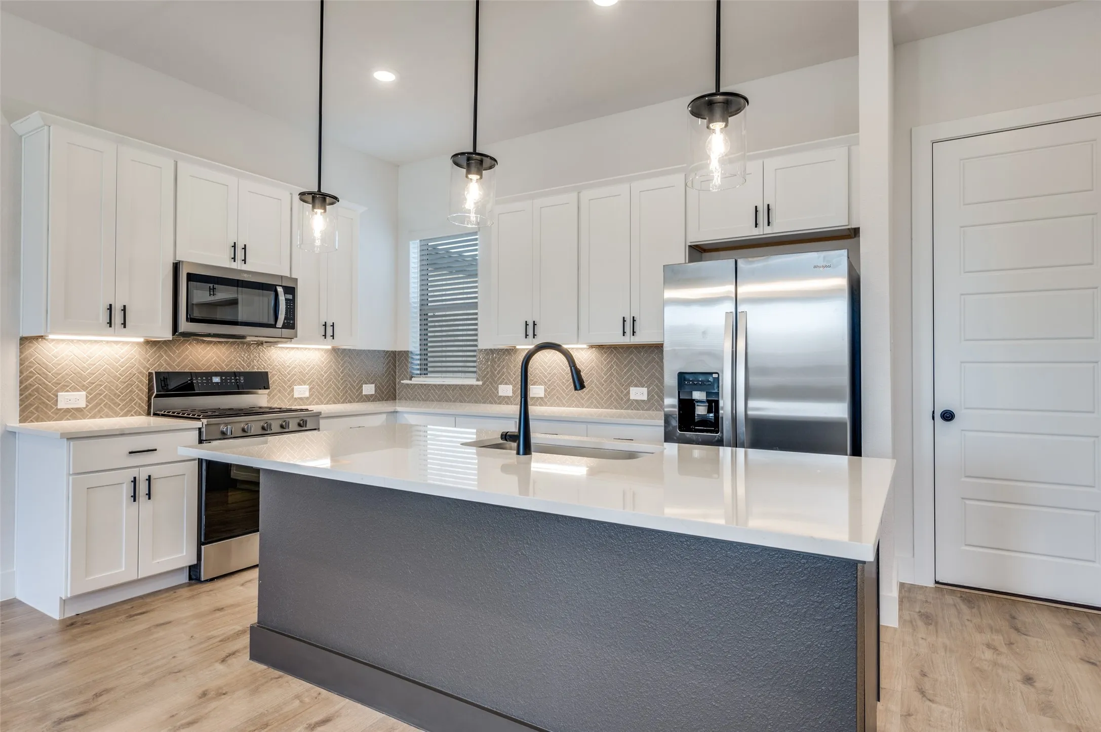 Bright, modern kitchen featuring crisp white cabinetry, quartz countertops, and a beautiful herringbone tile backsplash. The oversized island provides generous prep space and seating, complemented by stylish pendant lighting and a sleek black faucet. Stainless steel appliances, abundant cabinetry, under-cabinet lighting, and contemporary black hardware complete this beautifully designed and functional space.