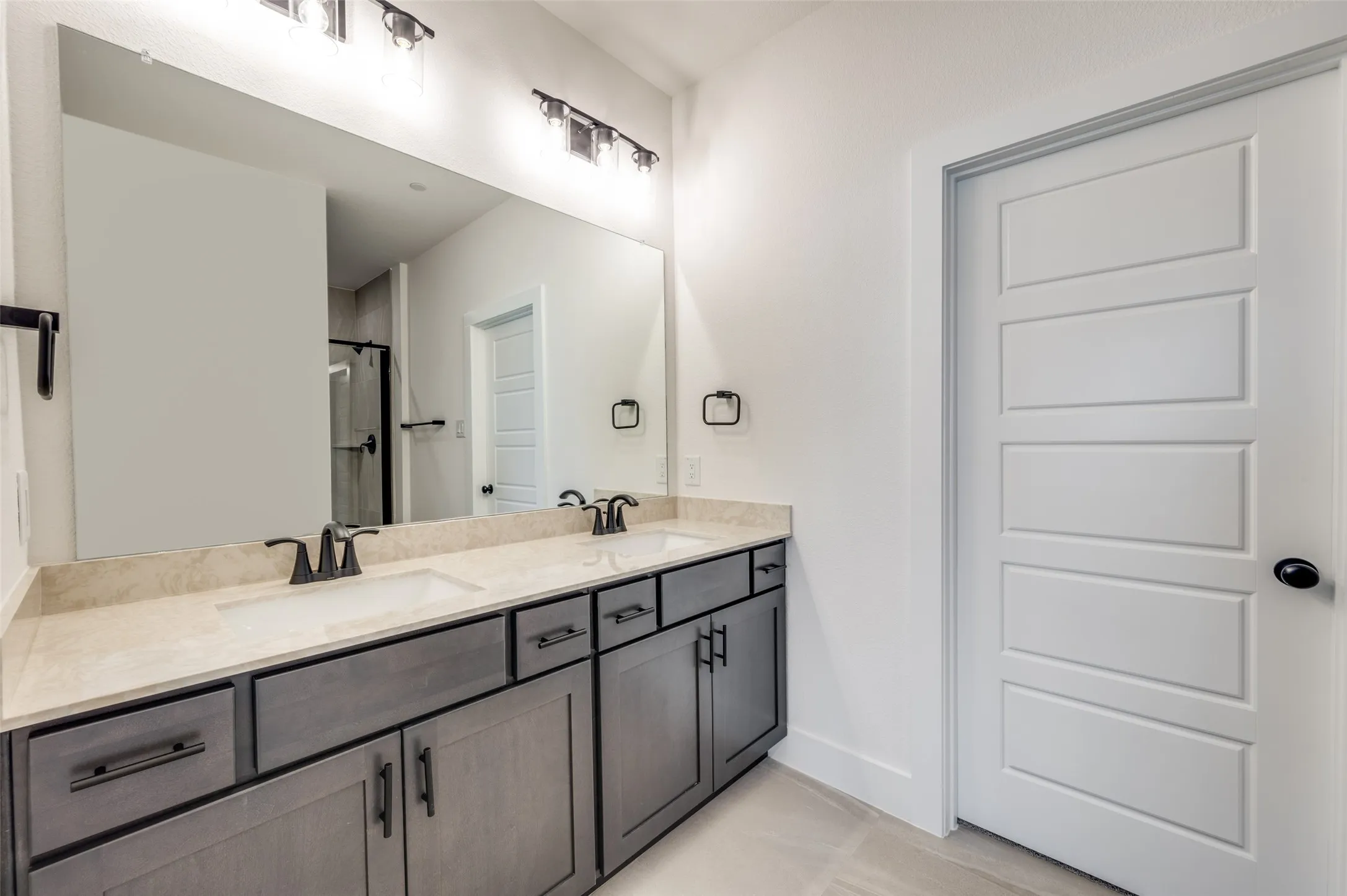 Spacious primary ensuite bath featuring a dual-sink vanity with abundant cabinetry, generous counter space, and a large wall-to-wall mirror. Sleek black fixtures and hardware add a modern touch, while the walk-in shower completes this stylish and functional private retreat.