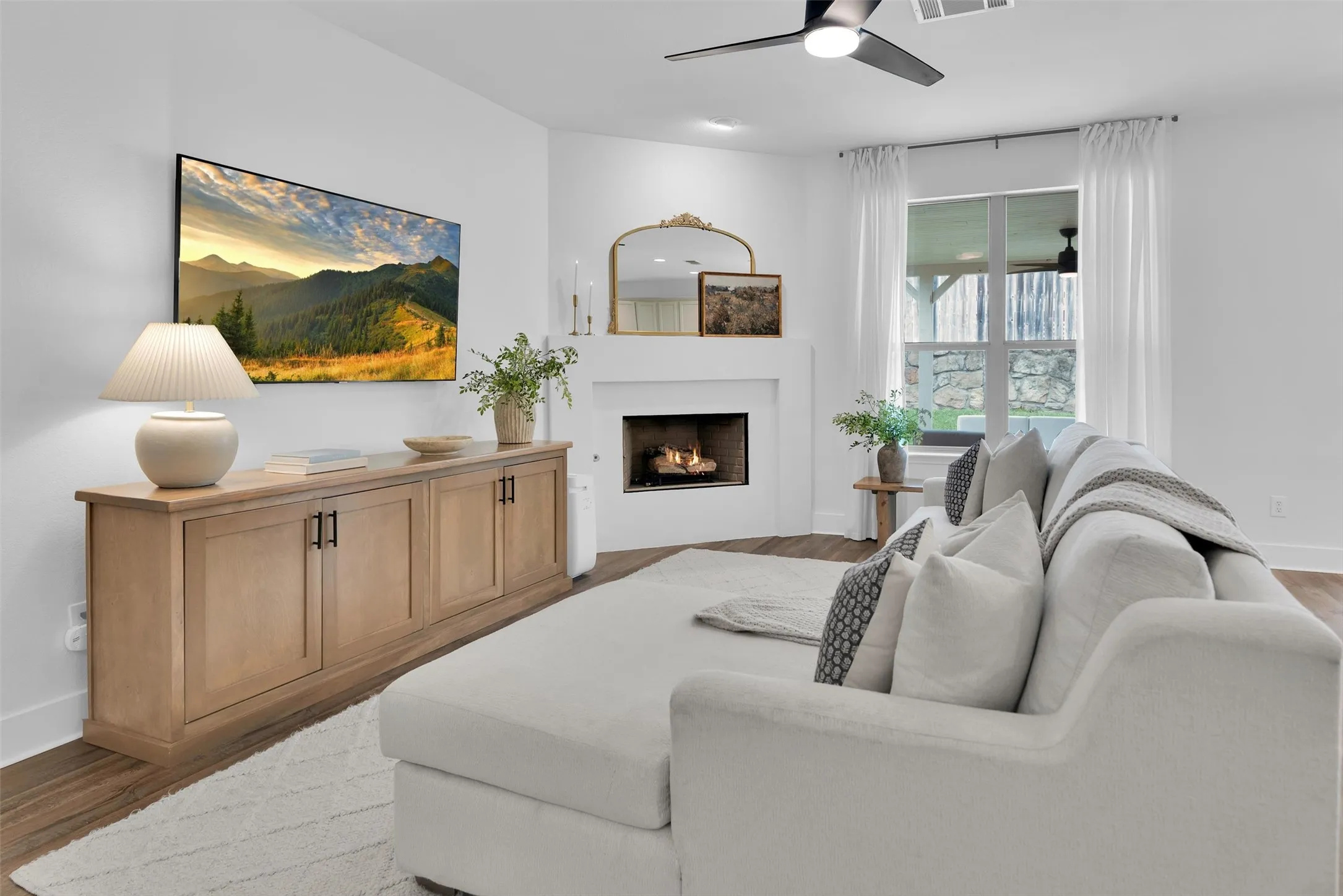 Hardwood-finish flooring, corner fireplace, ceiling fan, and large window with exterior stone elements