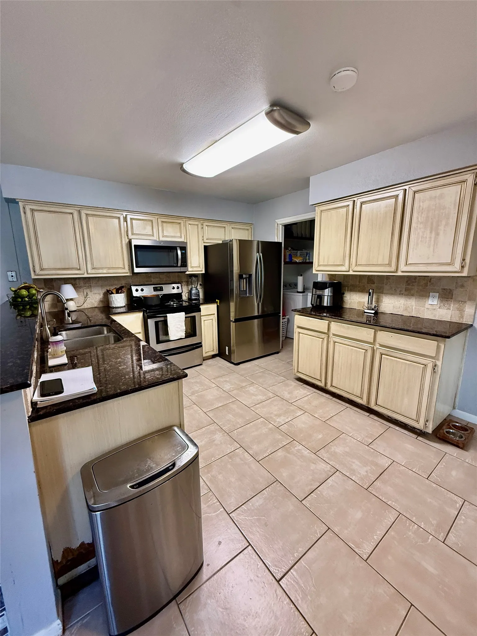Kitchen featuring light-toned cabinetry, stainless steel appliances, dark countertops, a tiled backsplash, and rectangular floor tiles