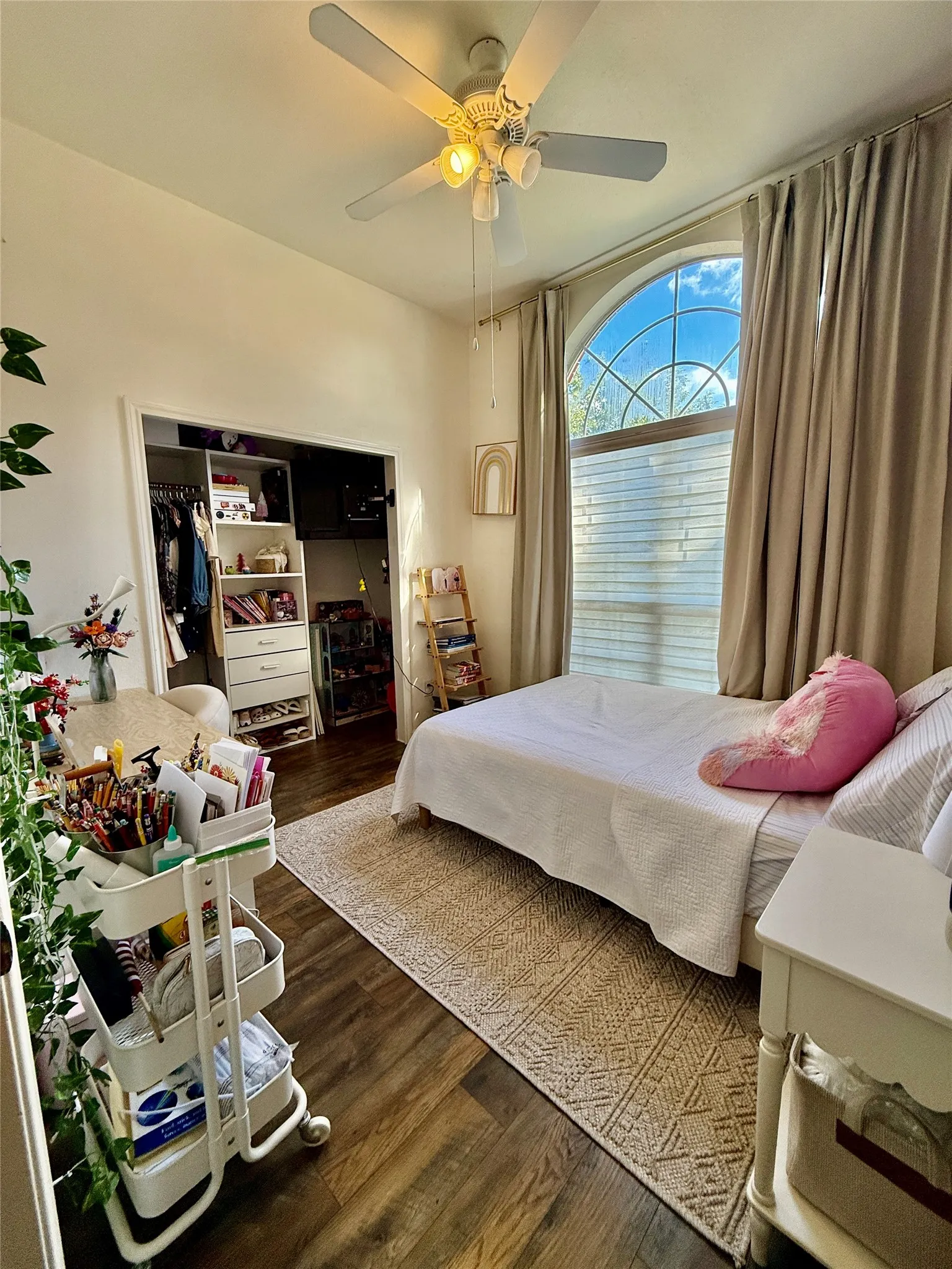 Arched window with decorative grilles, wood-finish flooring, ceiling fan with integrated lighting, and a closet with built-in shelving