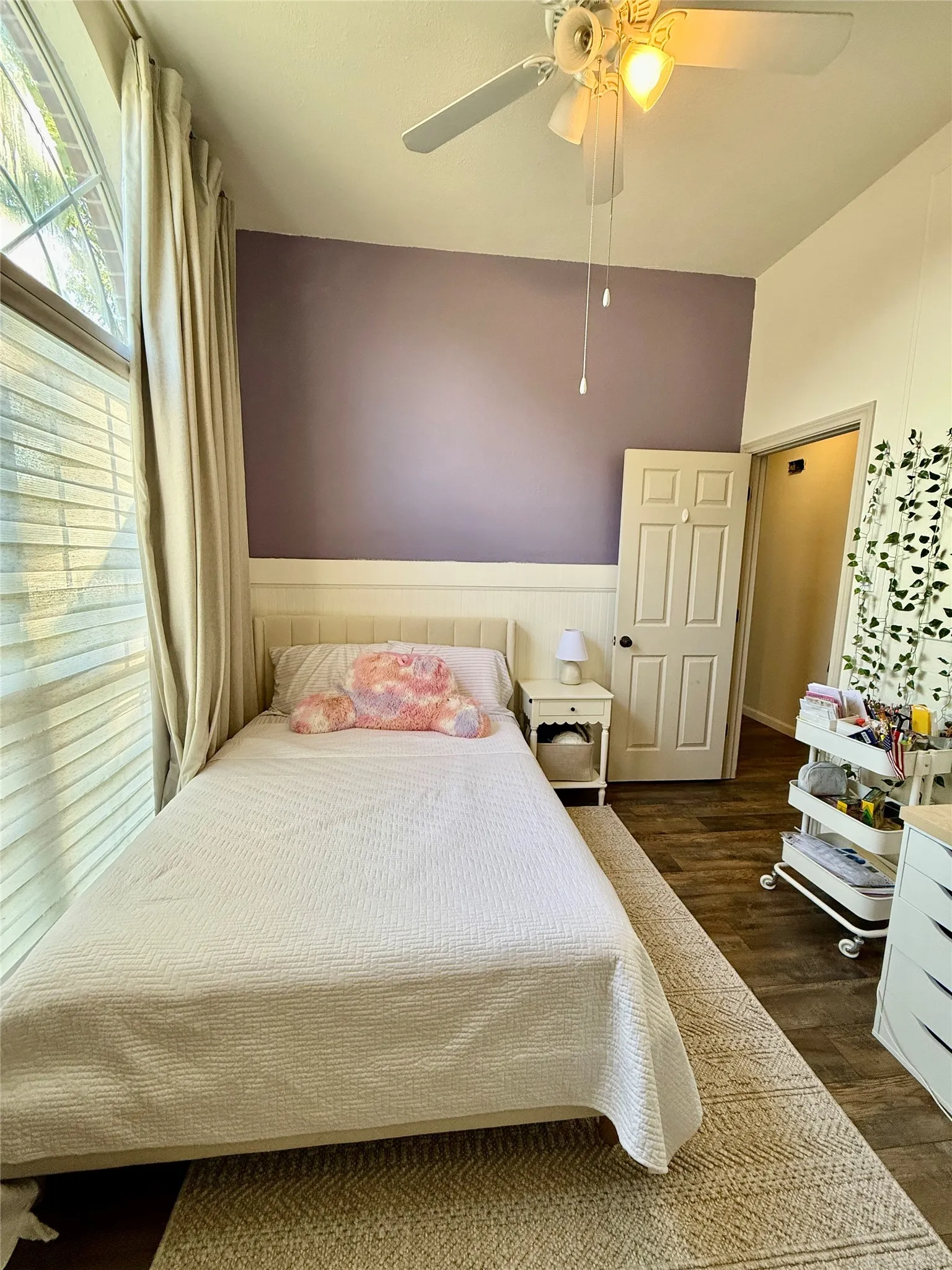 Angled window with horizontal blinds, overhead ceiling fan with lighting, wood-finish flooring, and white paneled door