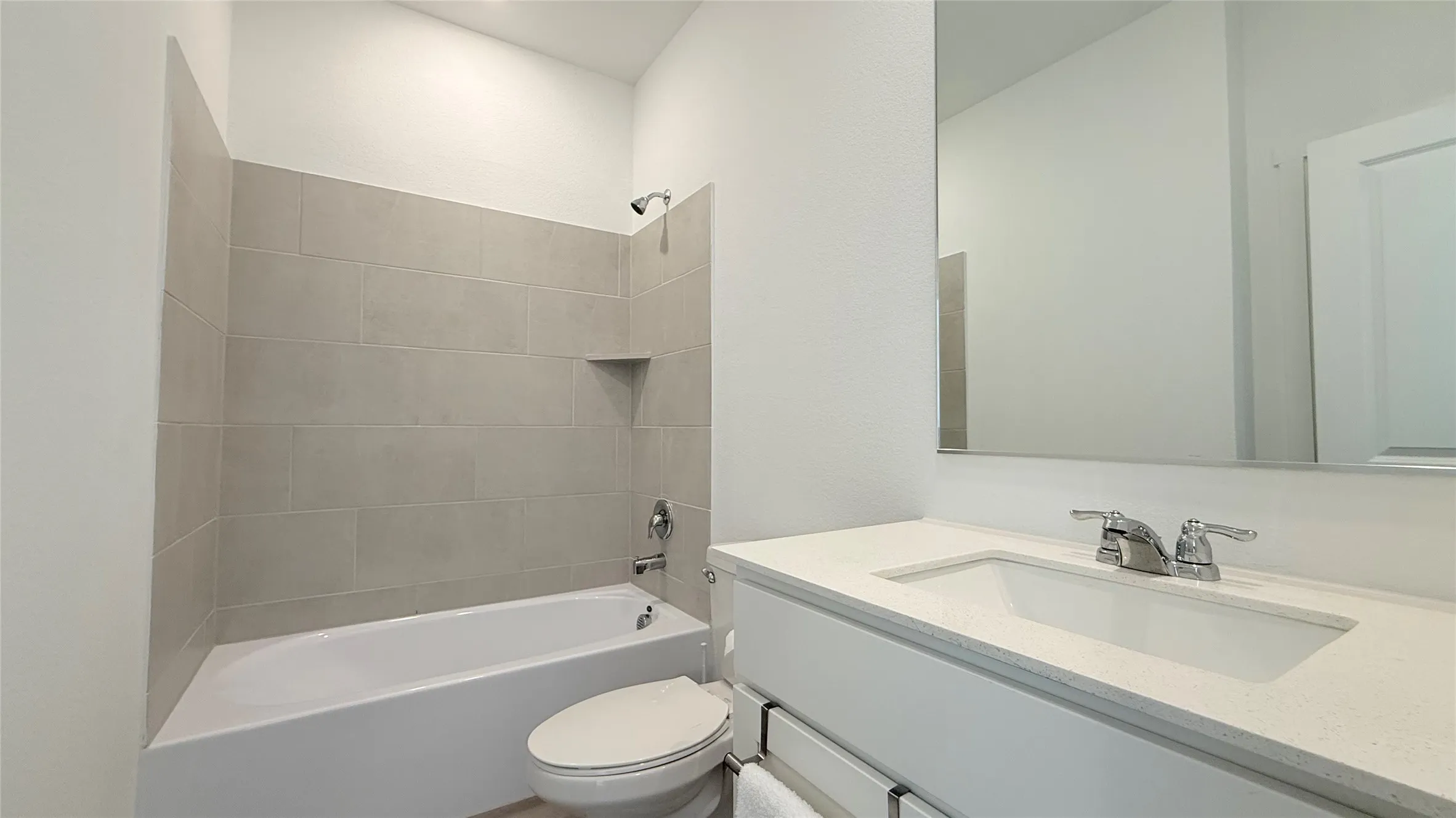 White vanity with an integrated sink and chrome faucet, large frameless mirror, and a tiled shower/tub combination with a recessed shelf