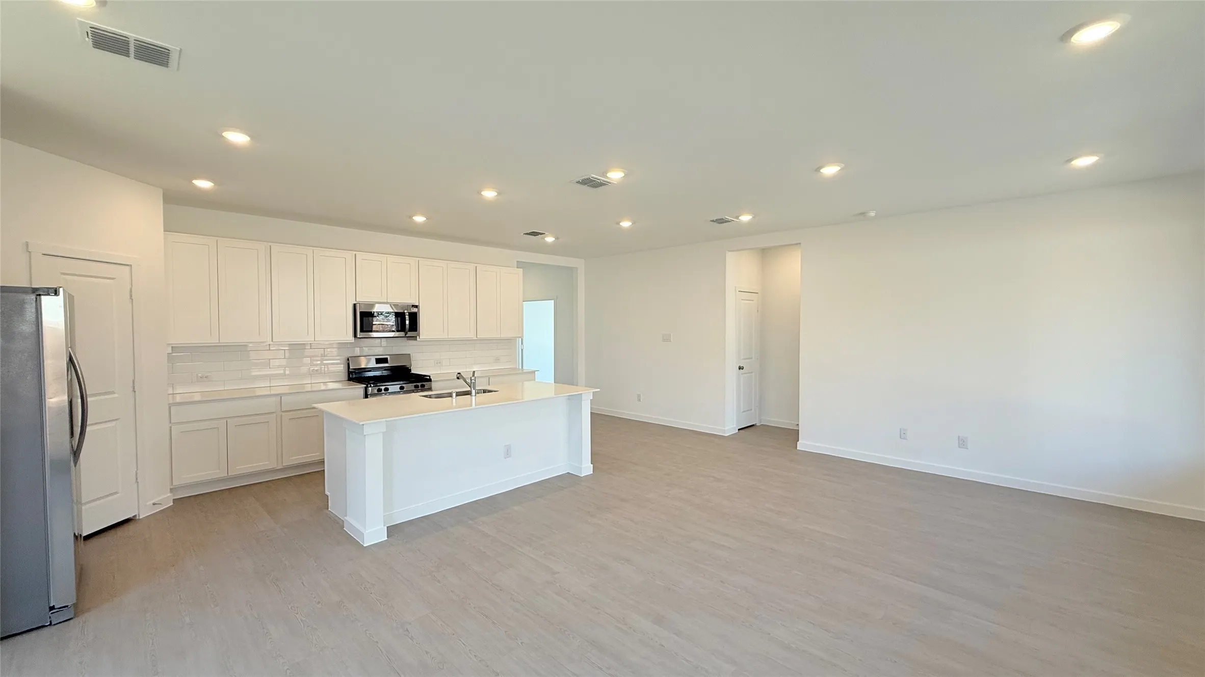 Spacious open-concept living area featuring wood-finish flooring, recessed lighting, white shaker-style cabinetry, and a kitchen island with an integrated sink
