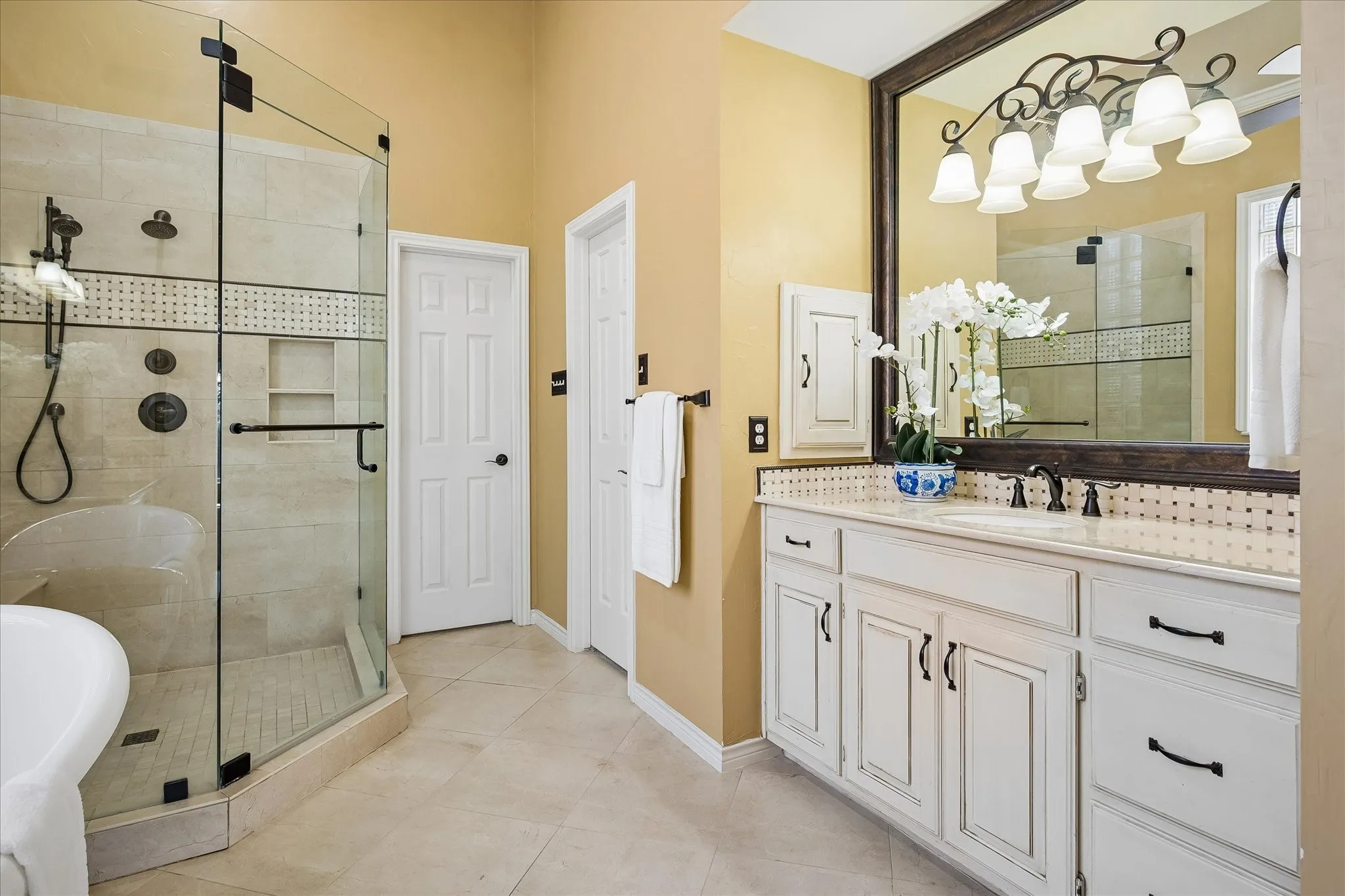 White vanity with bronze hardware, framed mirror, glass-enclosed shower with porcelain tile surround, built-in shower niche, and porcelain tile flooring.