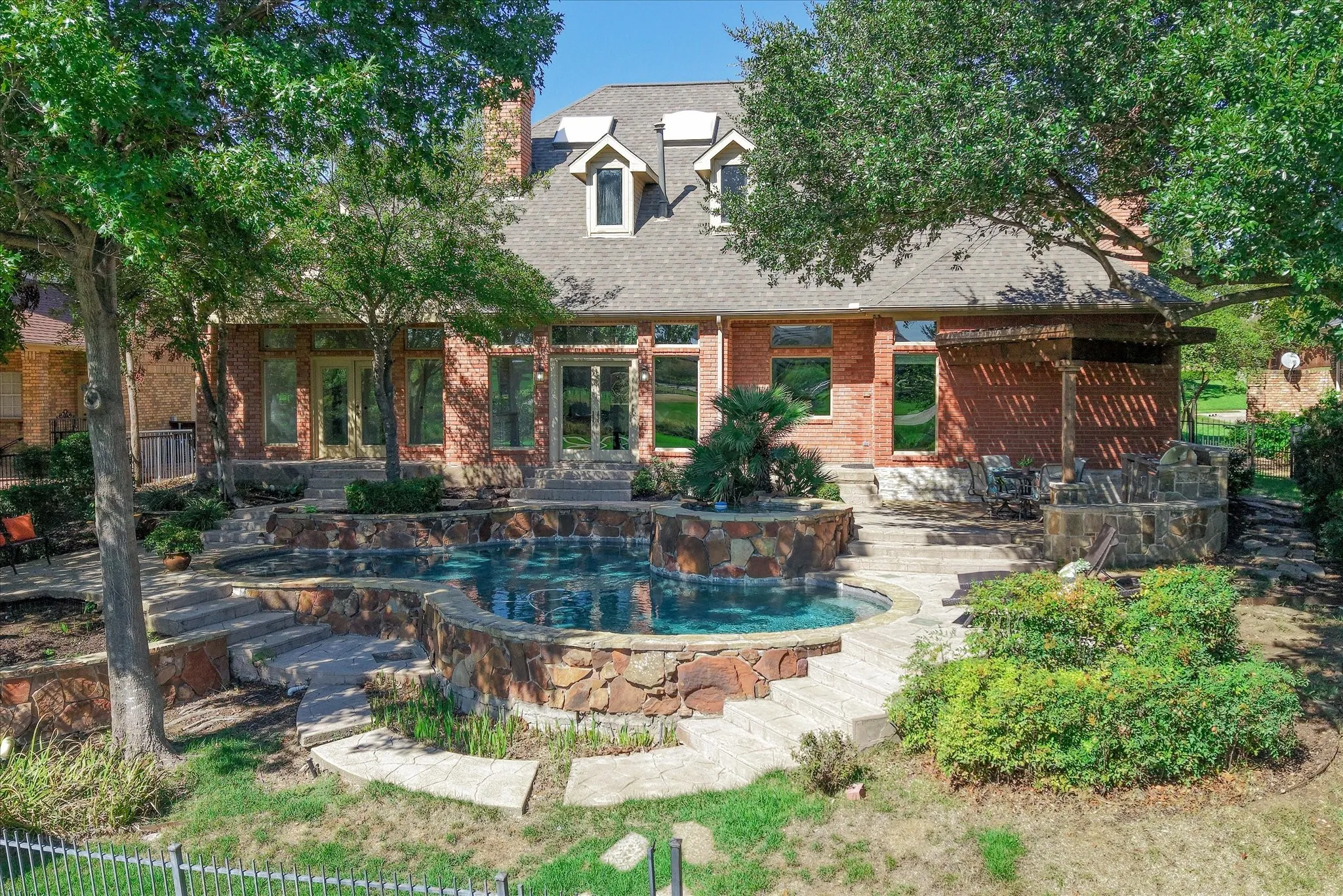Brick exterior with multiple windows, tiered swimming pool with integrated spa, stone retaining walls, and a stone patio