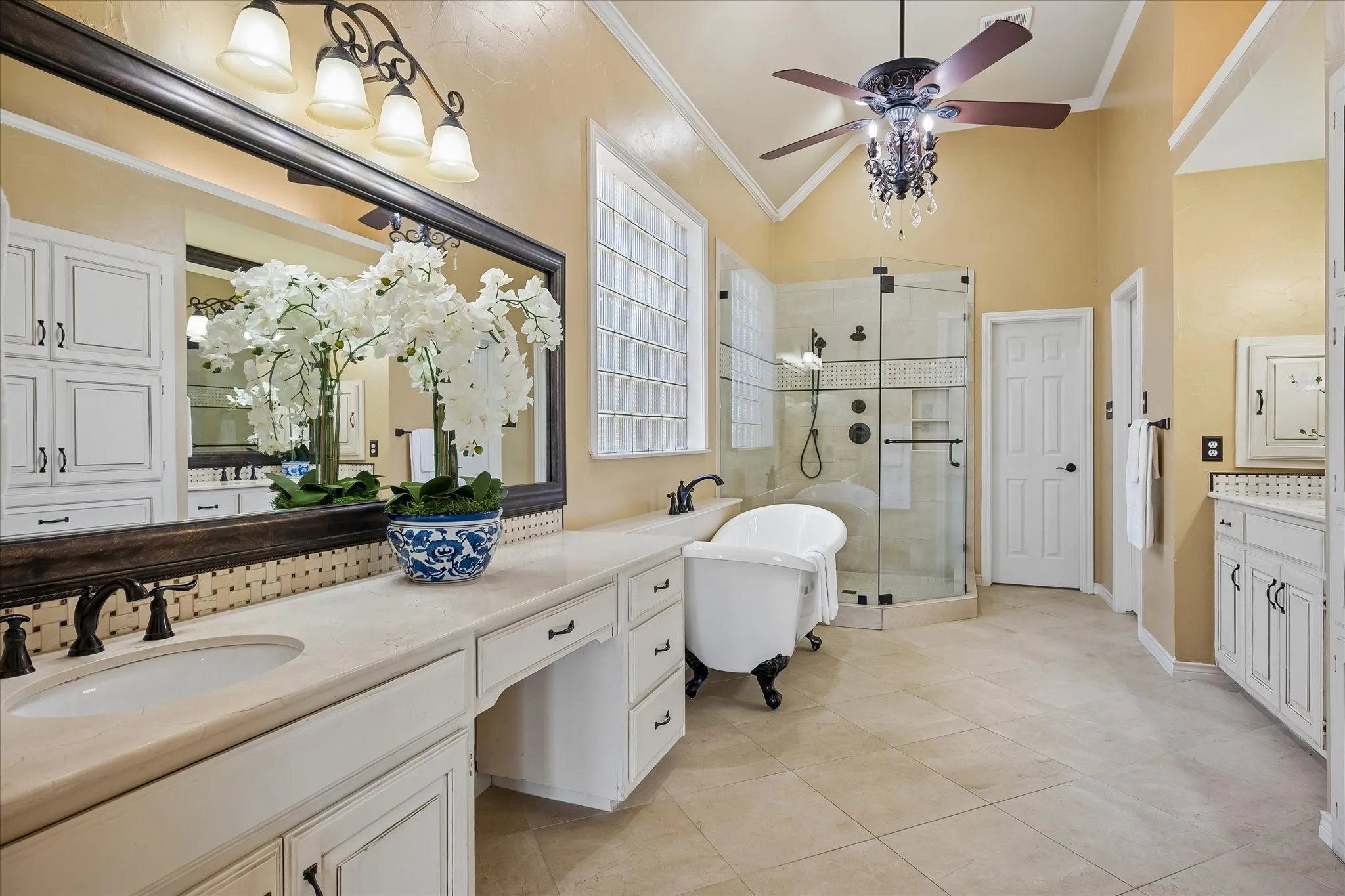 Spa-inspired primary bathroom featuring dual vanities with white cabinetry and marble countertops, bronze fixtures, a freestanding soaking tub, walk-in shower with frameless glass enclosure and tile surround, ceiling fan with integrated lighting, and neutral porcelain tile flooring