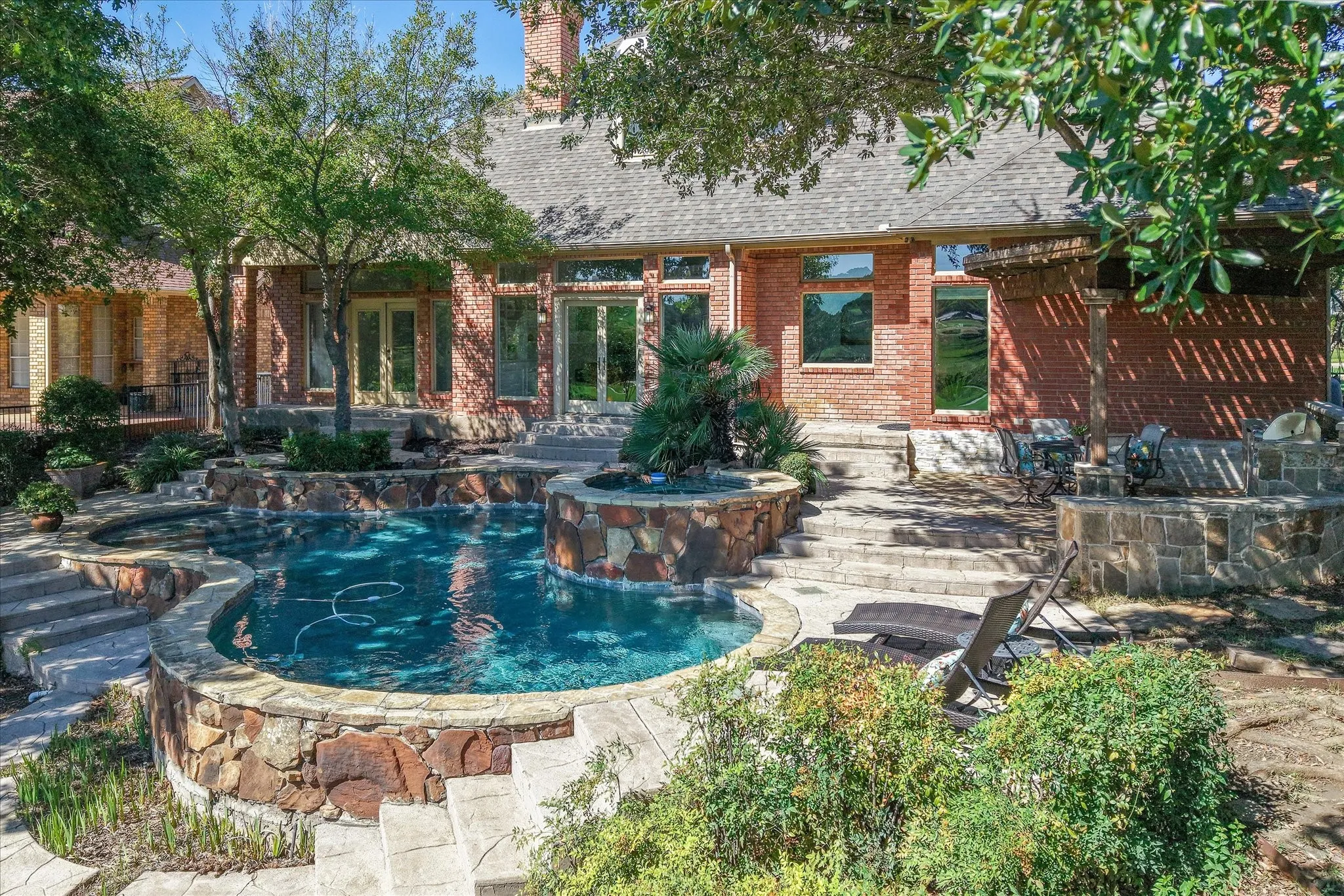 Brick exterior with multiple French doors, a multi-level pool with stone detailing, spa, and a stone-clad outdoor kitchen area