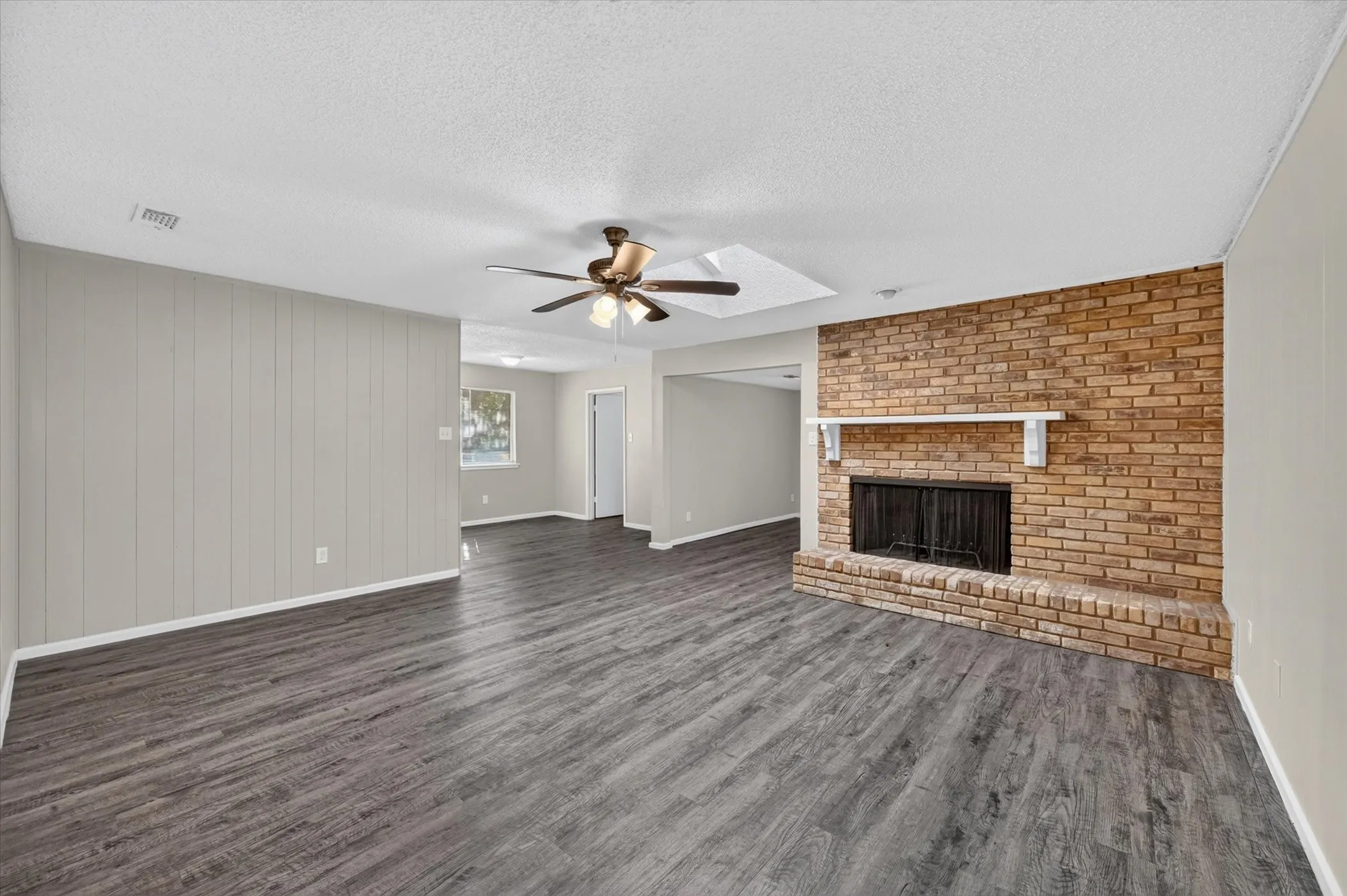 Spacious room featuring wood-finish flooring, a brick fireplace with a white mantel, and a ceiling fan with integrated lighting