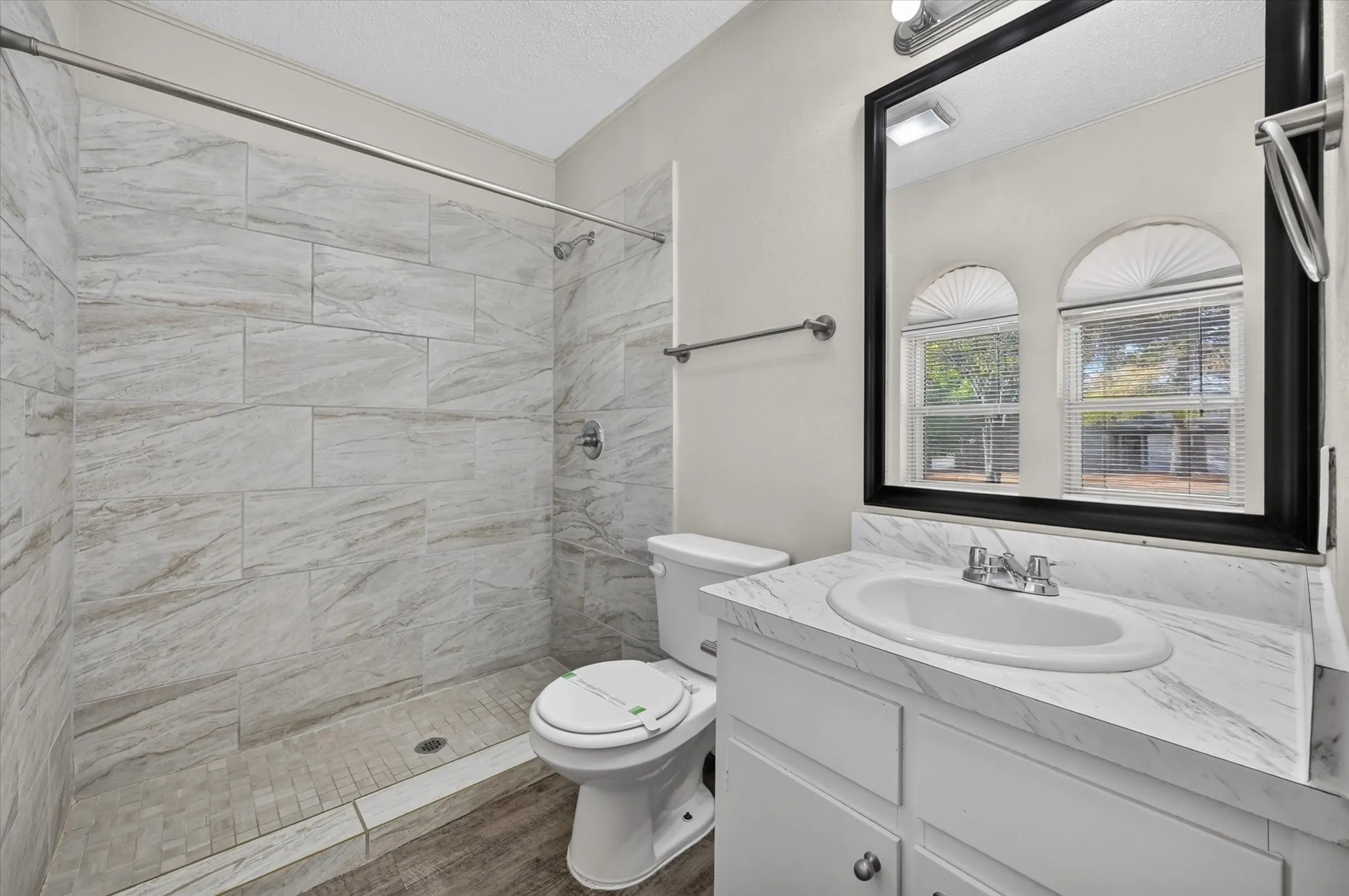 Walk-in shower with large format tile surround, white vanity with integrated sink, wood-finish flooring, and a framed mirror