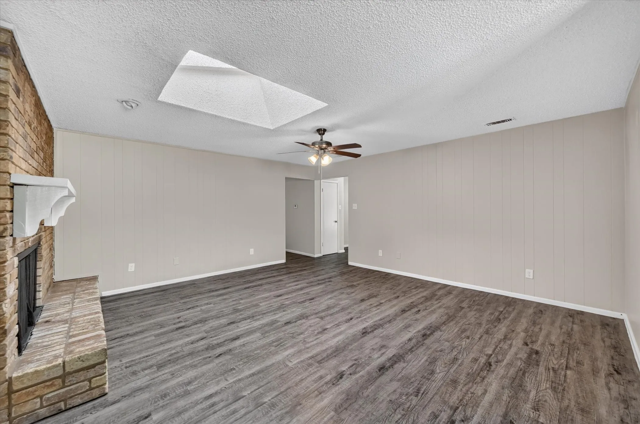 Living area featuring wood-finish flooring, a brick fireplace with a white mantel, vertical paneling, and a ceiling fan