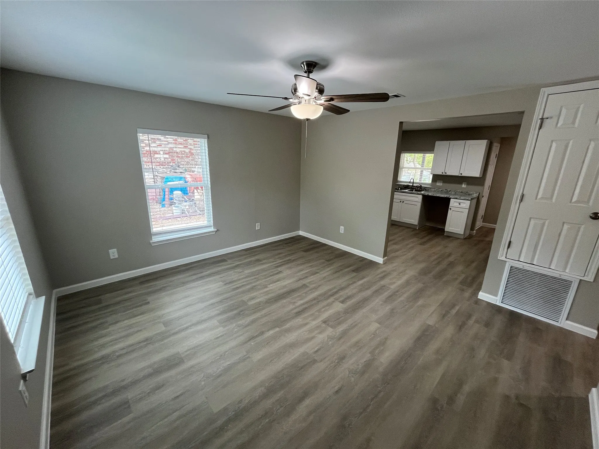 Gray wood-finish flooring, ceiling fan with integrated lighting, window with blinds, paneled door, and kitchen space featuring white cabinetry and light-toned countertops