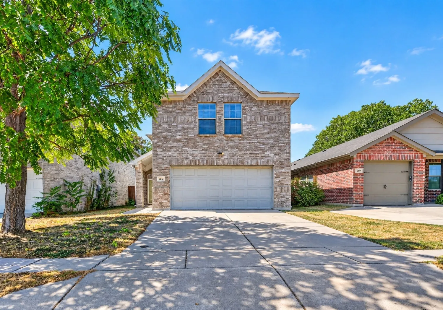 A well-maintained two-story single-family brick residence featuring a wide paved driveway, attached two-car garage, mature front lawn landscaping, and prominent entryway.