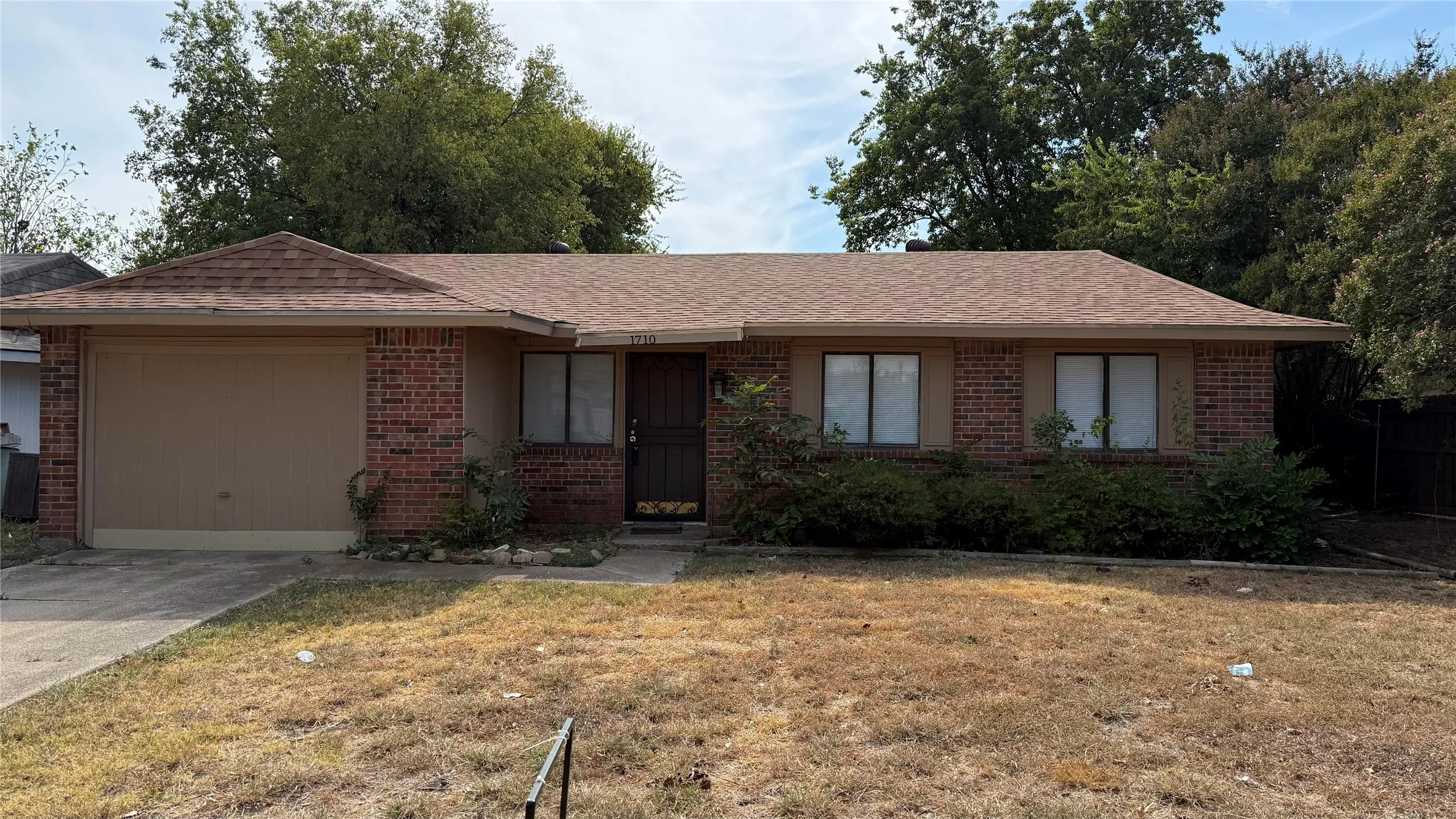 Brick facade with an attached garage, a brown shingle roof, and established front landscaping