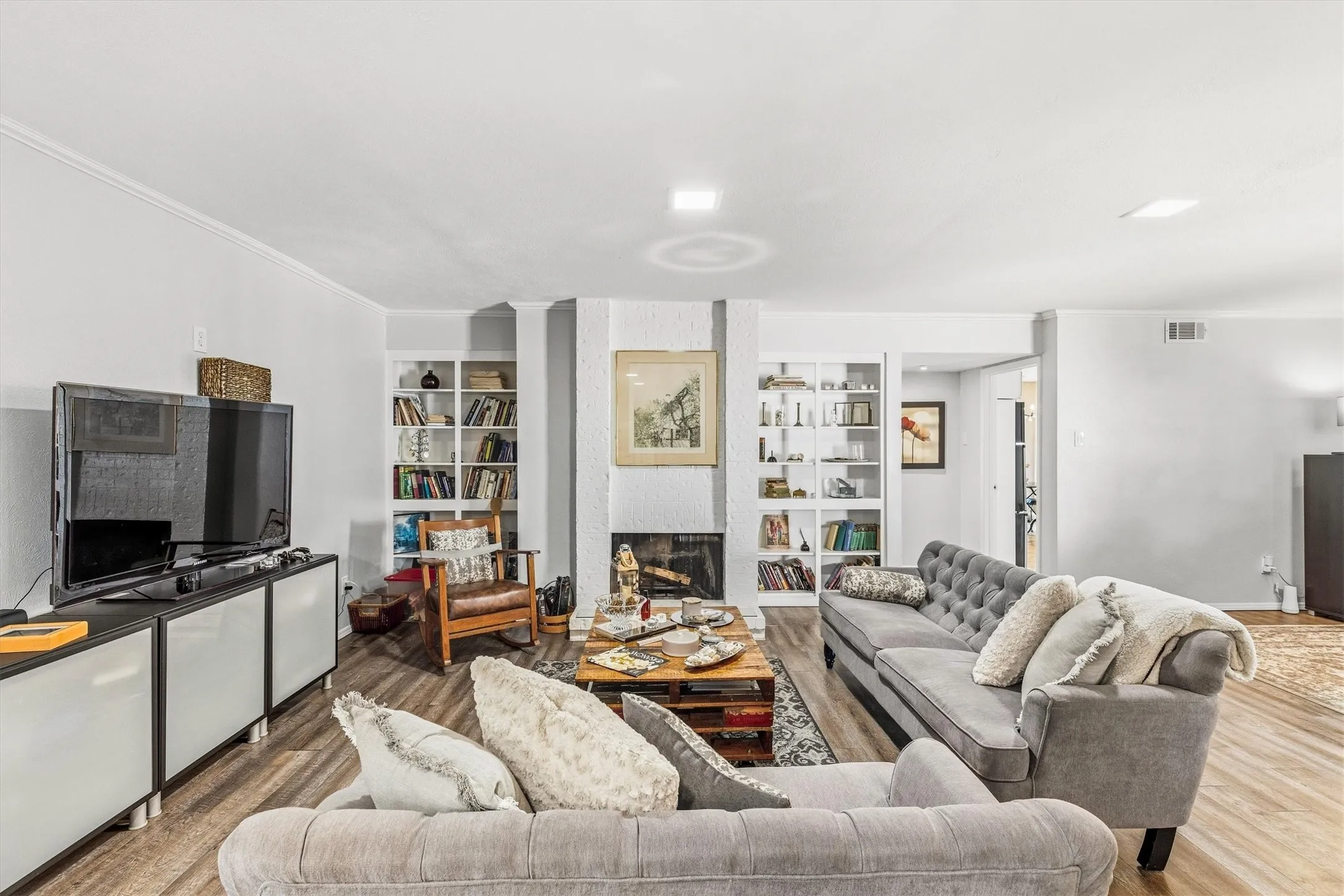 Wood-finish flooring throughout, featuring a white painted brick fireplace, built-in shelving, and recessed lighting
