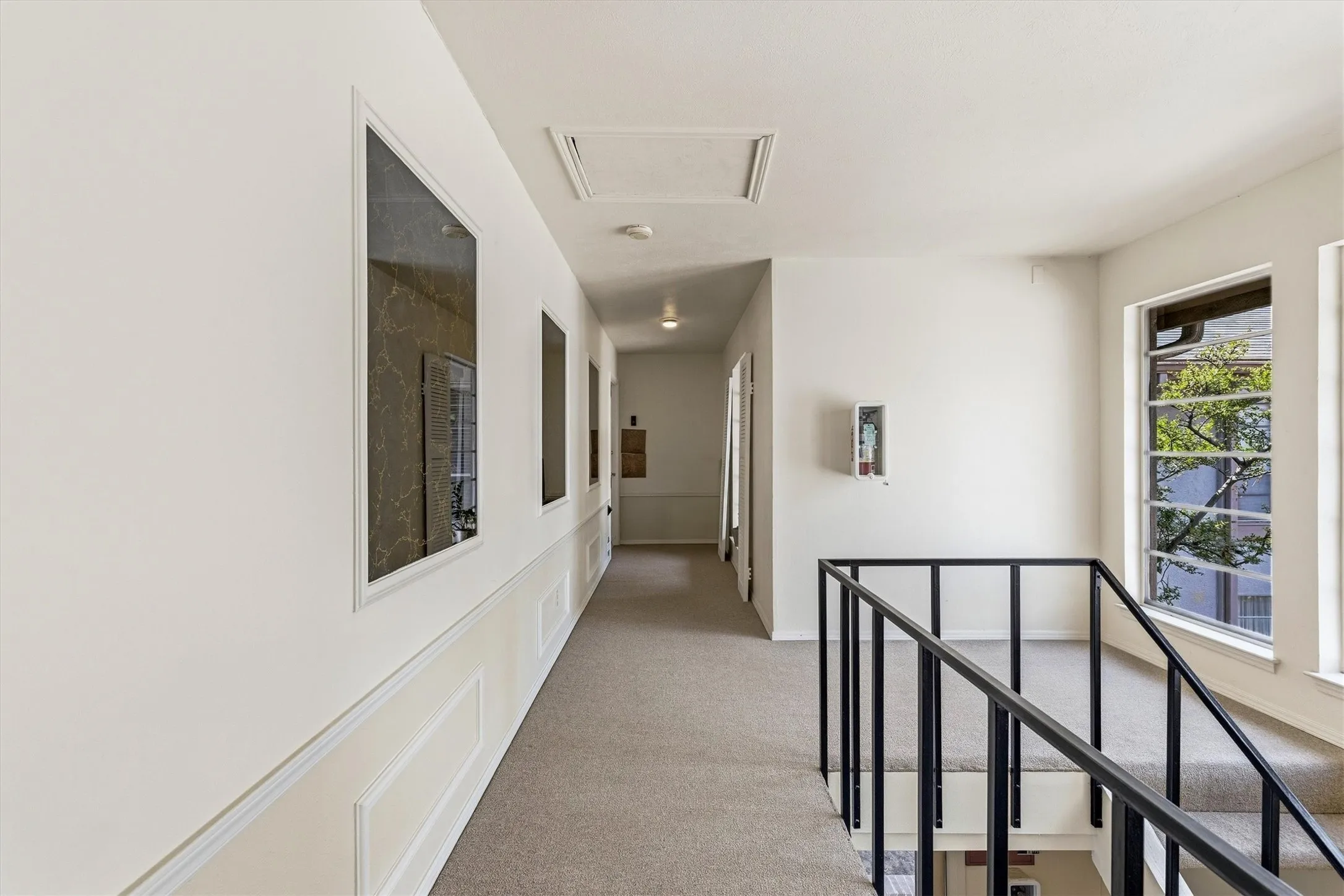 Hallway featuring light carpeting, wainscoting, and large windows with black metal grilles