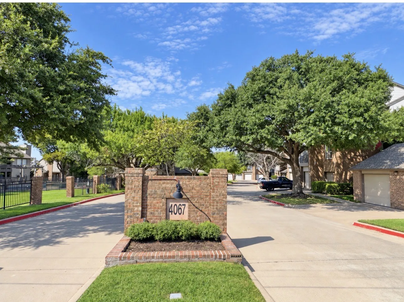 Brick entry monument with address signage, manicured landscaping, and mature trees