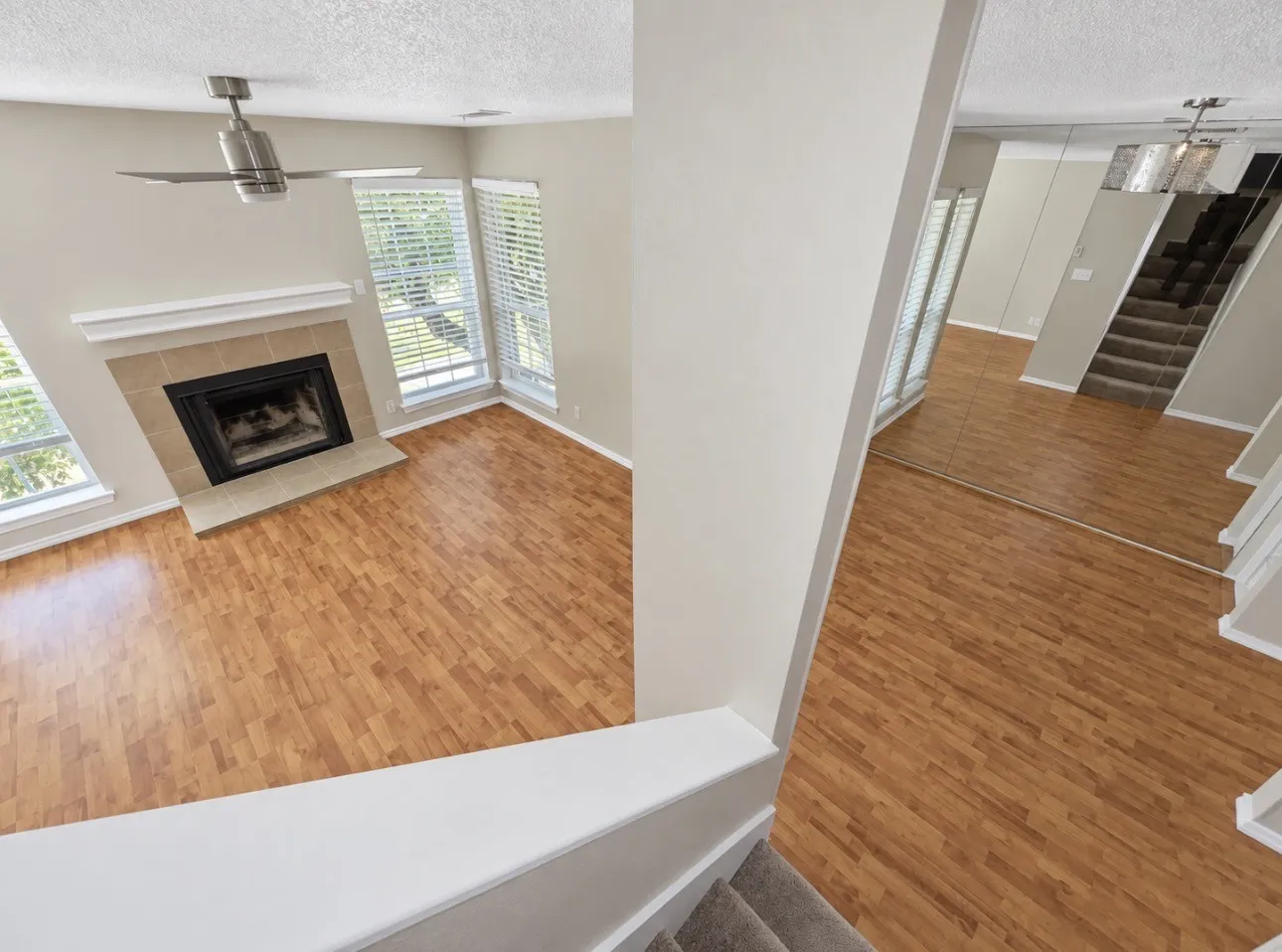 Wood-finish flooring, tiled fireplace with mantel, ceiling fan, multiple windows with blinds, mirrored wall accents, and carpeted staircase