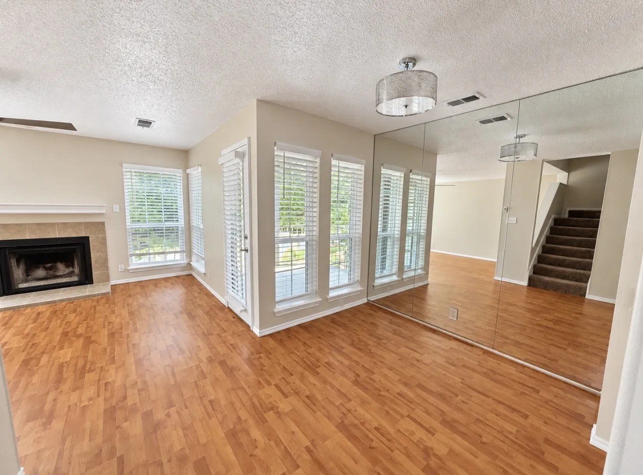 Wood-finish flooring throughout, fireplace with tile surround, multiple windows with blinds, mirrored wall feature, and a staircase with carpeted treads